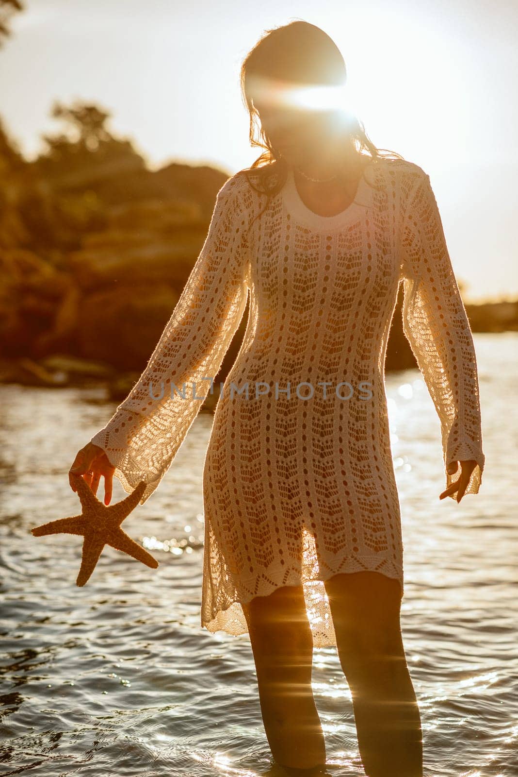 Beautiful young woman holding a starfish in her hands and enjoying it on the beach. The girl is standing in backlight. Her dress is glowing in the sun.