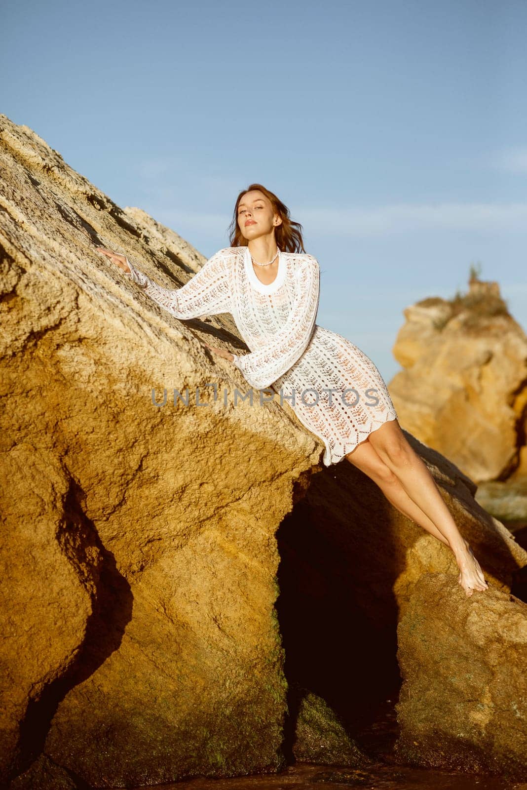 Beautiful young woman posing on a large rock on the sea coast. girl sitting on a rock in a white knitted dress, against the background of a rock