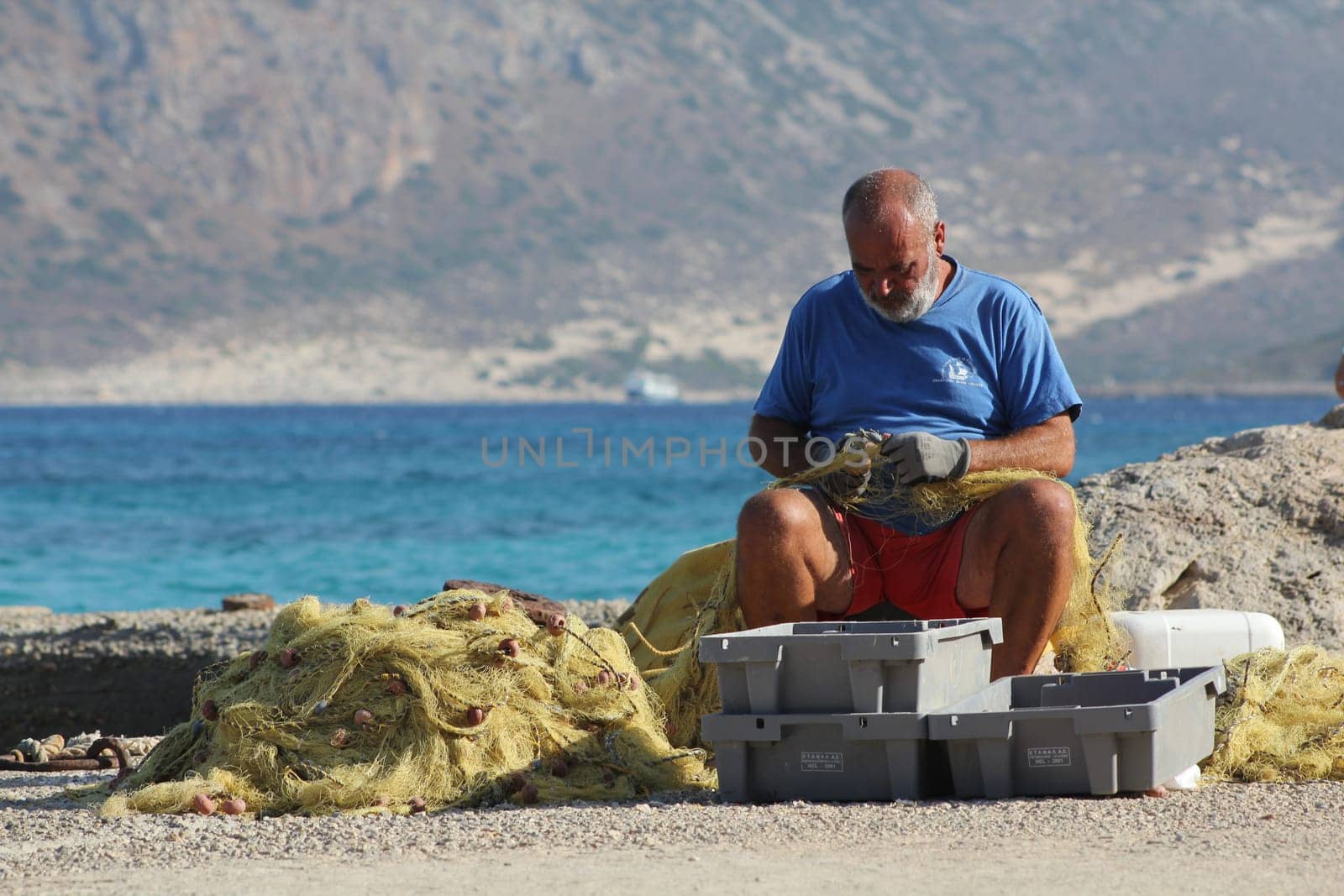 An adult man repairs tackle and fishing nets on the beach against a backdrop of mountains in Balos Bay, Greece