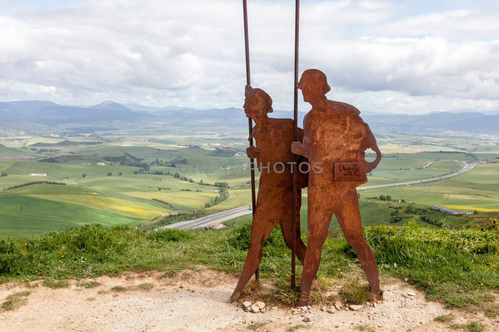 On the Camino de Santiago, metal sculptures of pilgrims walk toward stunning hills under a cloudy sky.