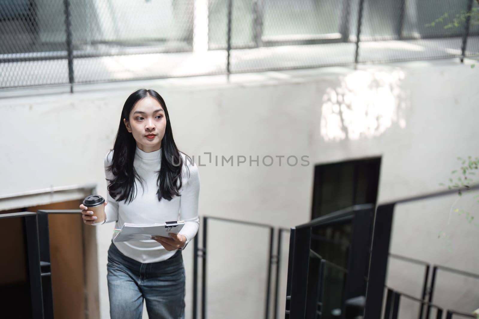 A young professional woman is ascending stairs while holding documents and a coffee cup, demonstrating focus and determination in her work.