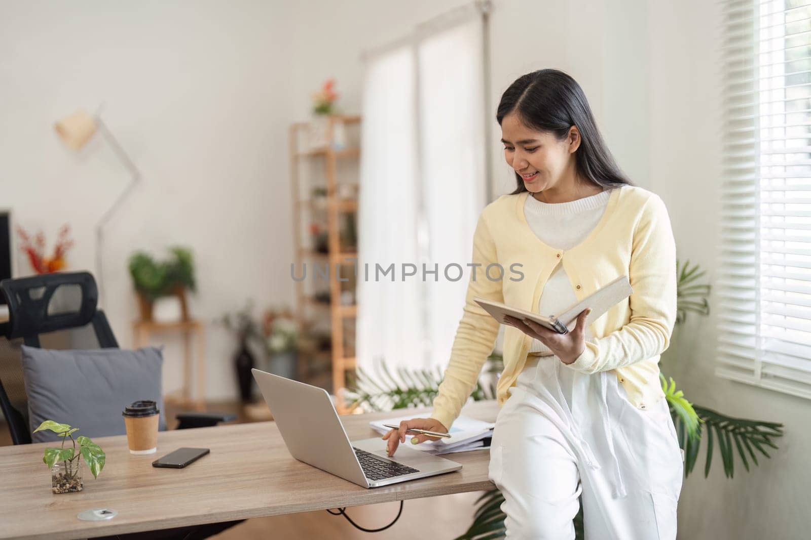 A young woman stands confidently by her desk, engaging with her laptop and notes, showcasing a productive home office setup.