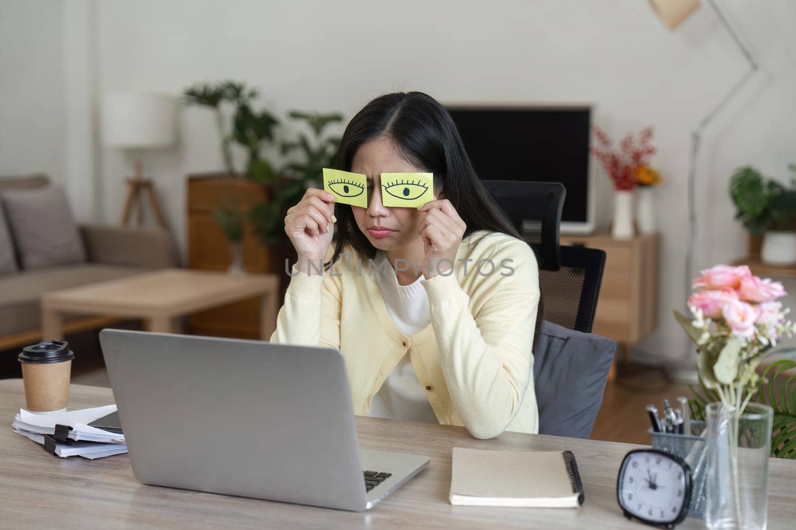 A woman in a cozy home office uses sticky notes with eyes drawn on them to combat fatigue while working on her laptop.