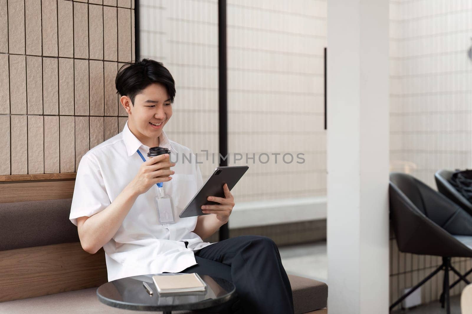 A young Asian male office worker smiles as he interacts with a tablet in a contemporary workspace, savoring his coffee.