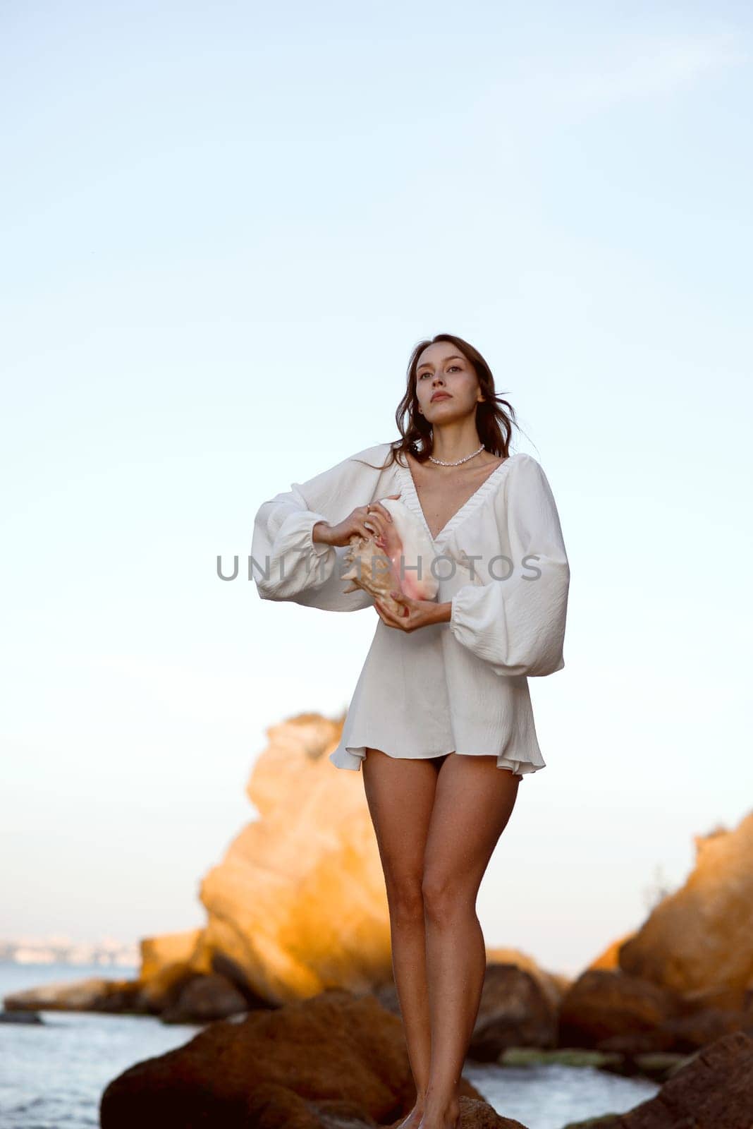 Young woman in summer white dress holds big shell in her hands. She is standing in sea, on beach. Behind her are rocks. Tourist trip abroad vacation, travel concept.