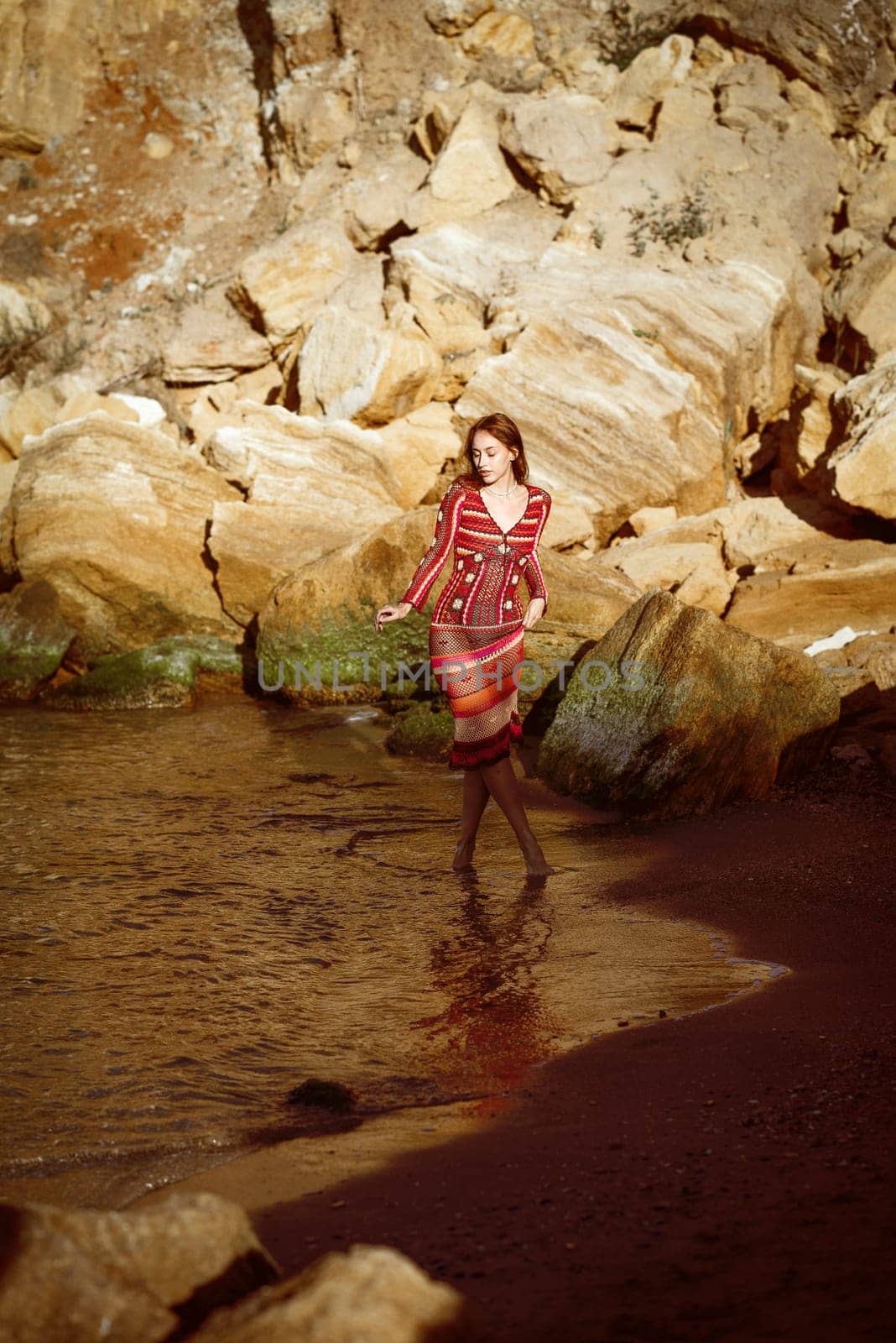 She is standing in the sea among huge rocks. Handmade dress, crocheted to fit. Boho style dress. She is walking along the seashore, general plan.