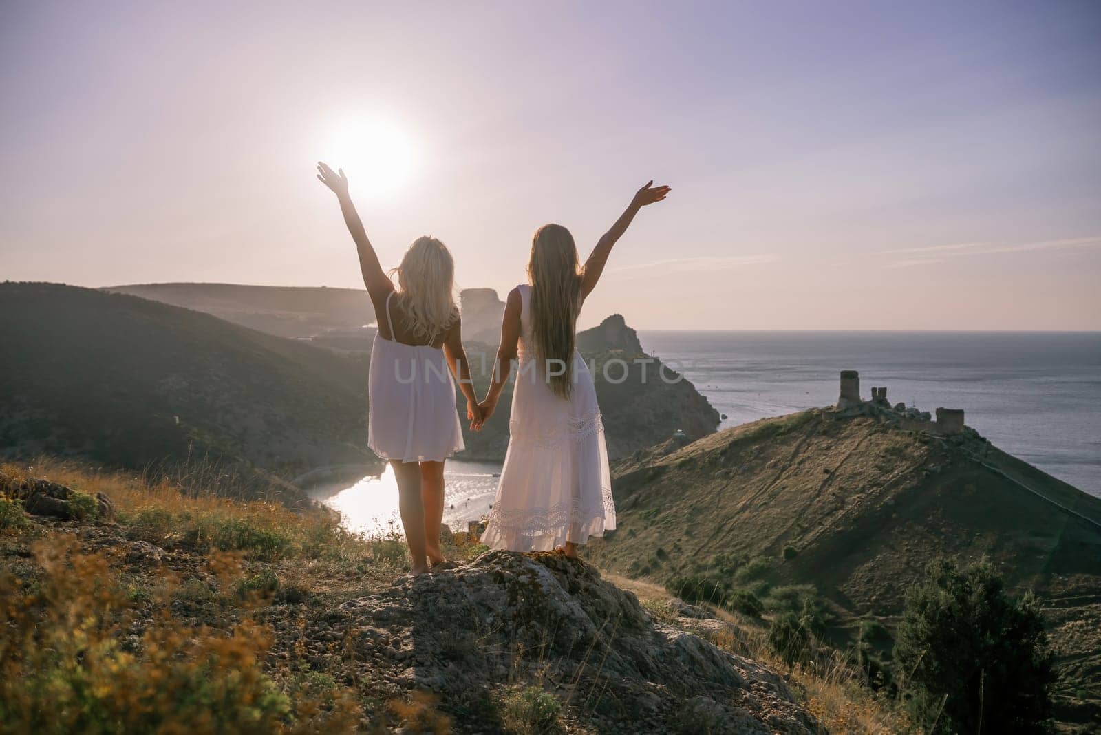 Two women are standing on a hill overlooking the ocean. They are holding hands and looking out at the water. The scene is peaceful and serene, with the sun shining brightly in the background