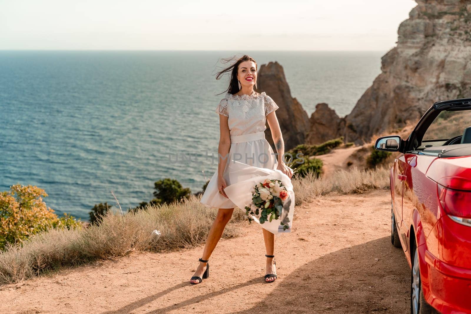 A woman in a white dress is standing next to a red convertible car. She is holding a bouquet of flowers and she is in a happy mood. The scene suggests a romantic or special occasion