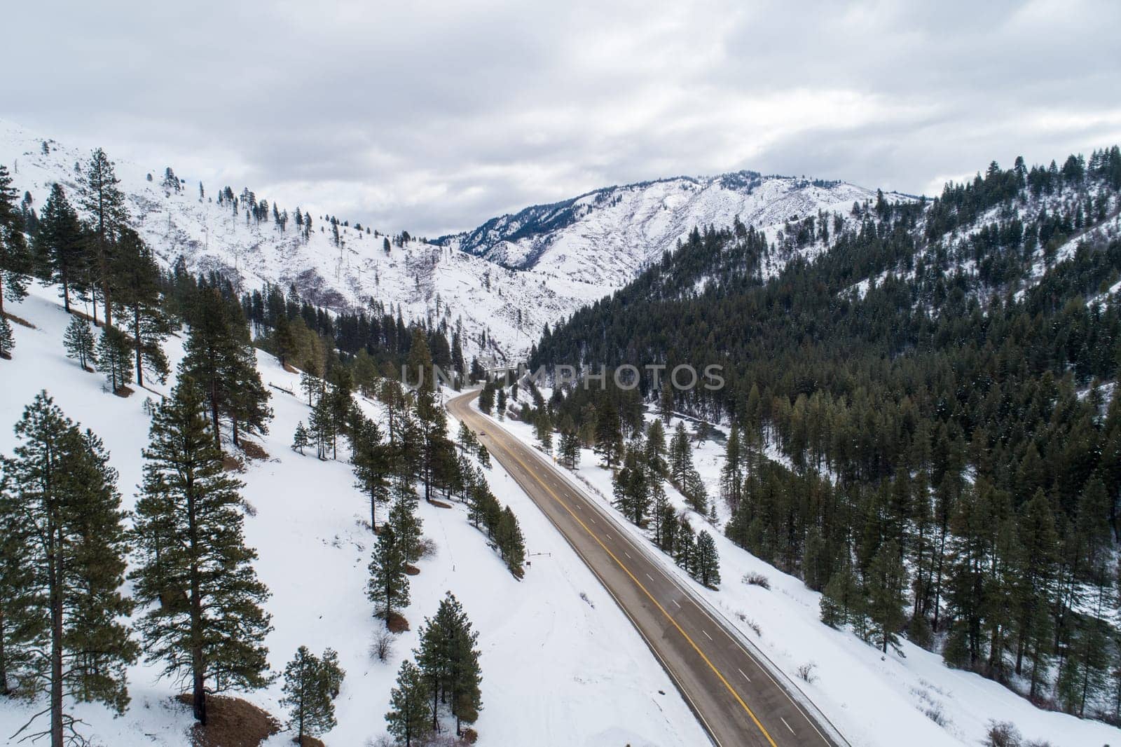 A snowy mountain range with a highway running through it. The sky is cloudy and the trees are bare