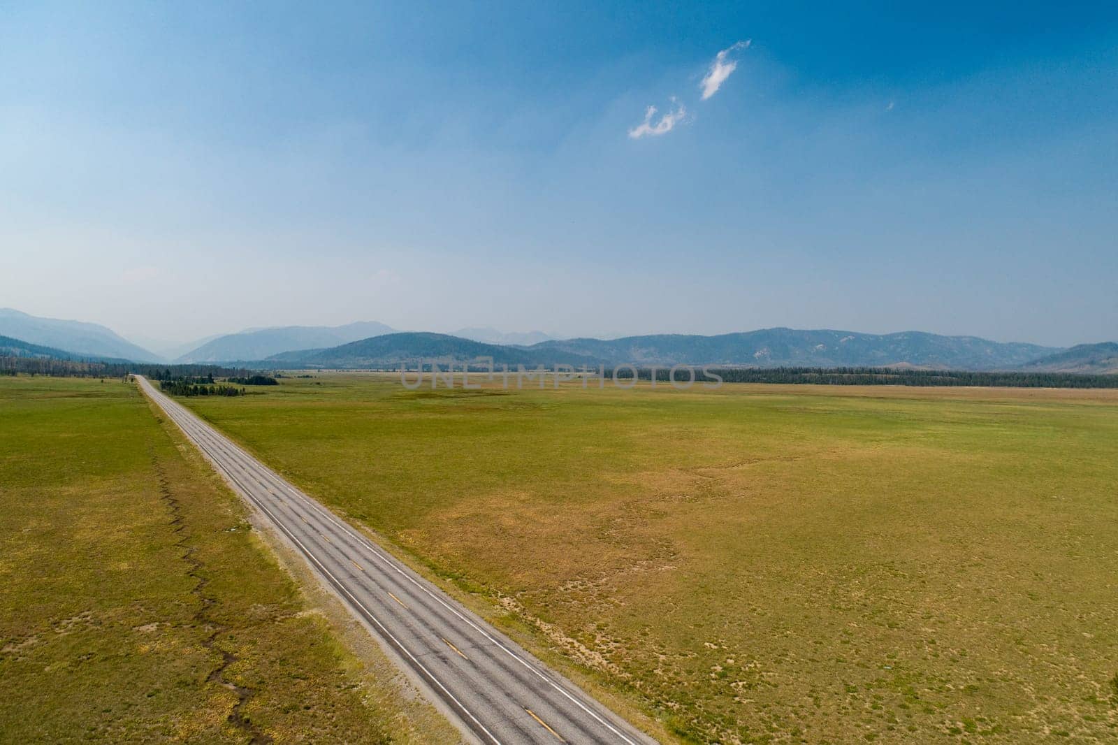 A long road stretches across a vast, empty field. The sky is clear and blue, and the sun is shining brightly. The scene is peaceful and serene, with no signs of human activity