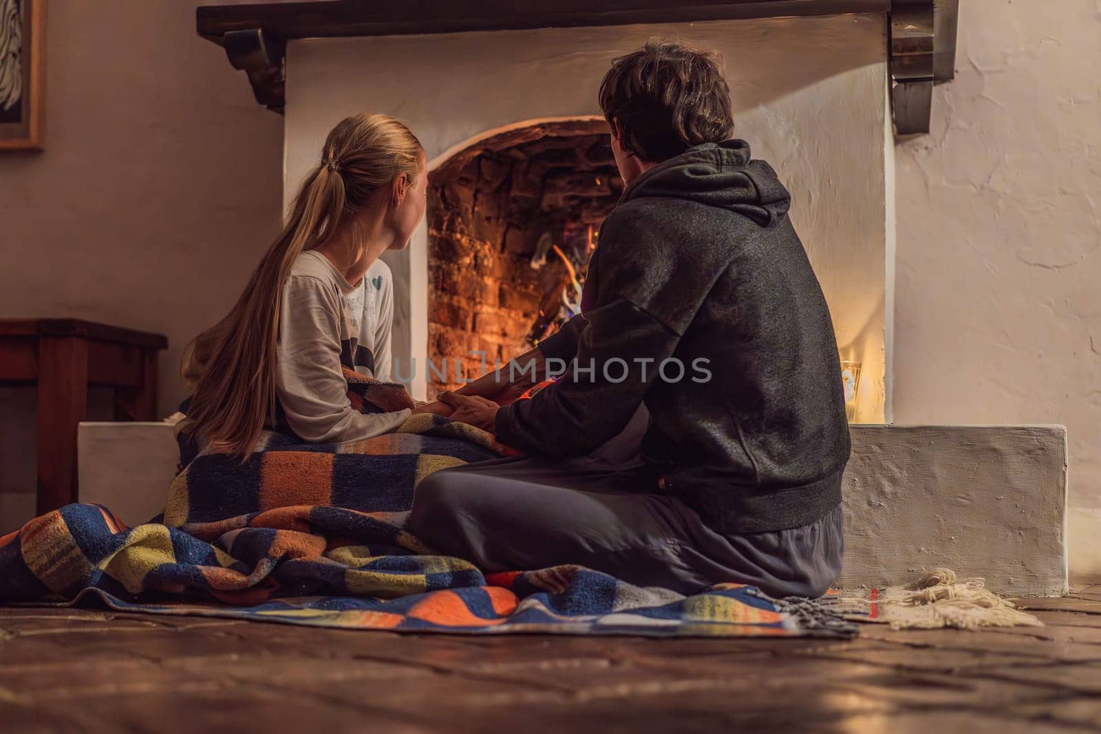 Family bonding by the fireplace during cozy winter evening. Warm and peaceful family moment indoors. Togetherness, love, and relaxation concept. A warm and inviting scene featuring a lit fireplace with glowing firewood and candles. A colorful traditional rug and pillow lie on the floor, creating a cozy atmosphere. Home, relaxation, and rustic comfort concept.