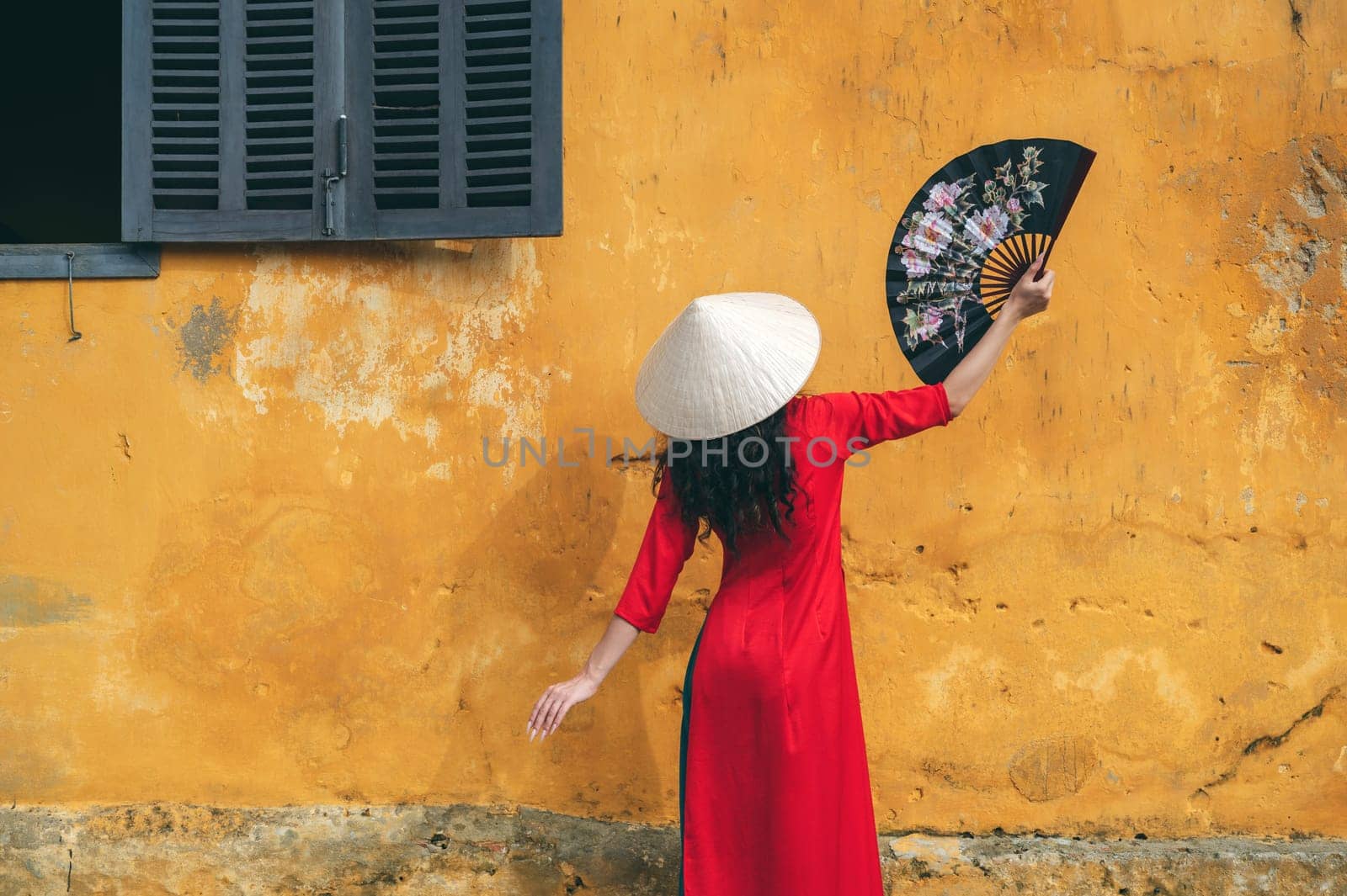 back of woman traveler tourist in traditional vietnamese hat and red dress with fan on old town street in Hoi An in Vietnam in Asia