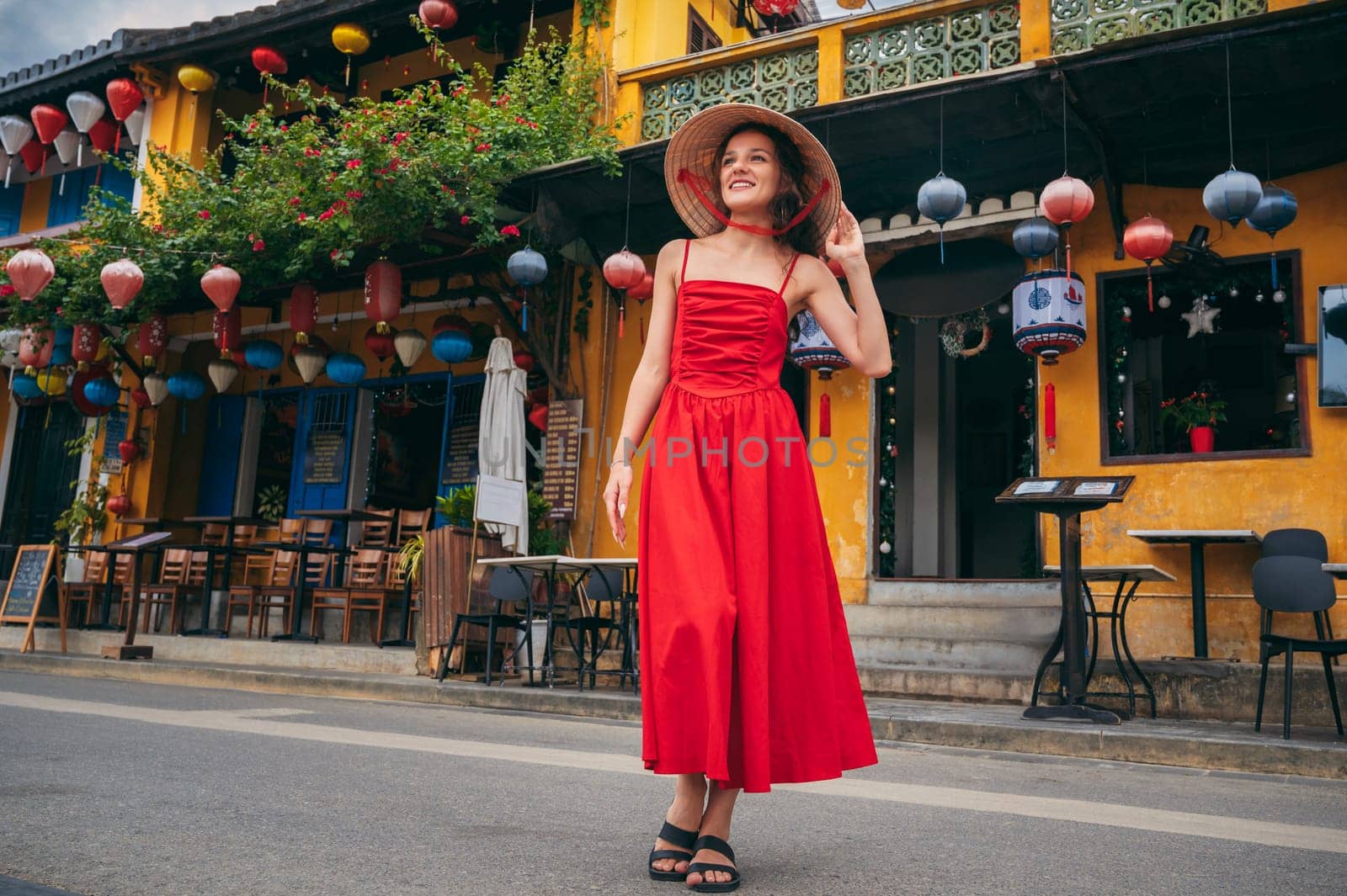 happy young female tourist traveler in traditional Vietnamese hat and red dress on street in old town in Hoi An city in Vietnam in Asia