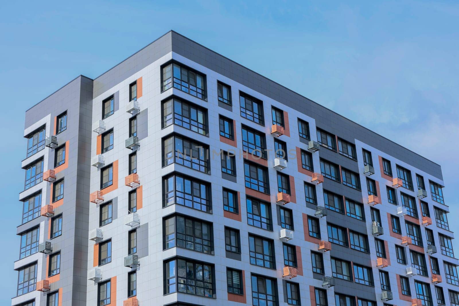 The concept of modern architecture, a residential complex, an apartment building for living. Close-up view from the corner of a multi-storey, apartment building in gray and white tones with orange inserts of modern architecture with large windows, panoramic balconies and baskets for air conditioning against the blue sky. High quality photo