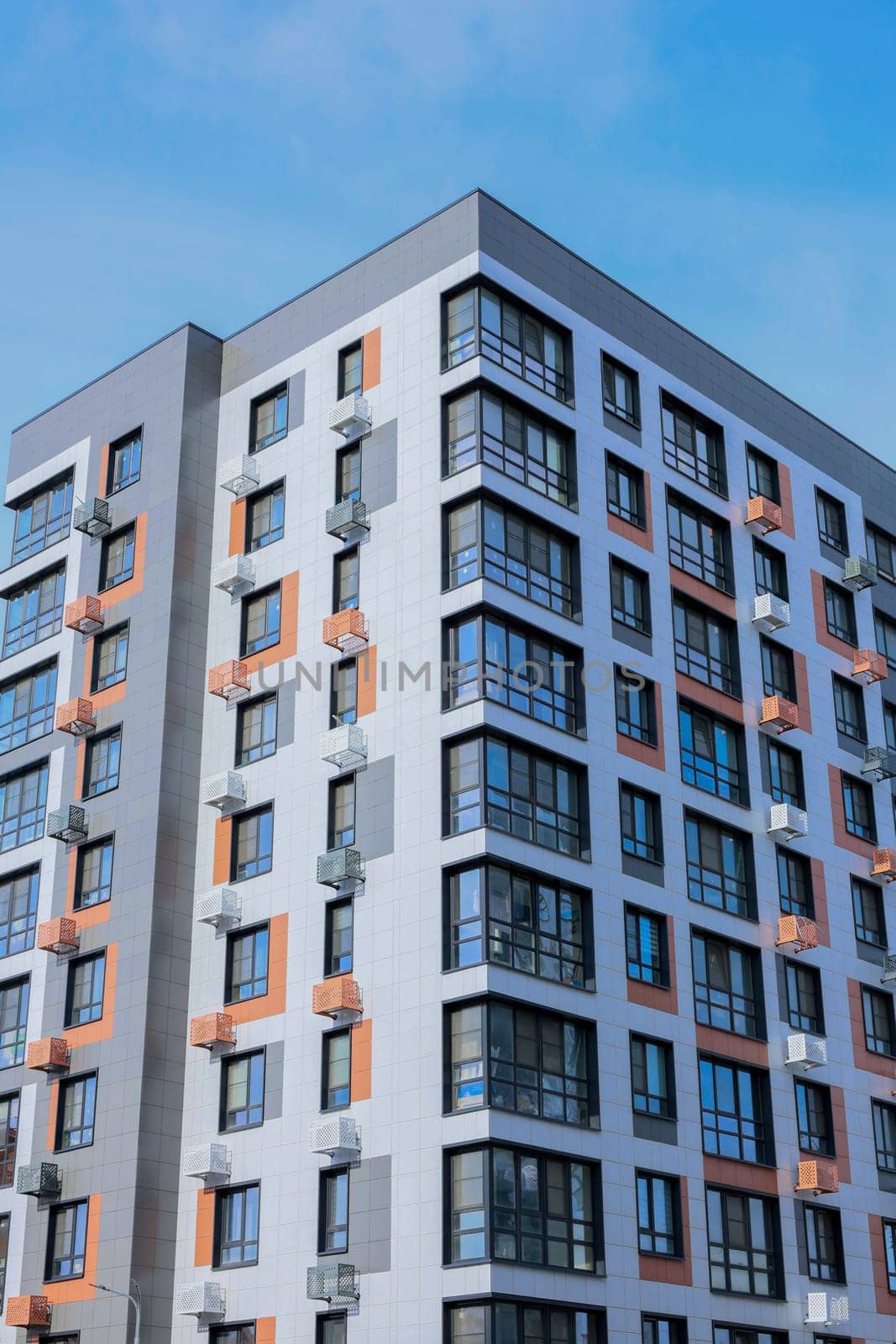 The concept of modern architecture, a residential complex, an apartment building for living. Close-up of a multi-storey, apartment building in gray and white tones with orange inserts of modern architecture with large windows, panoramic balconies and baskets for air conditioning against the background of a blue sky. High quality photo