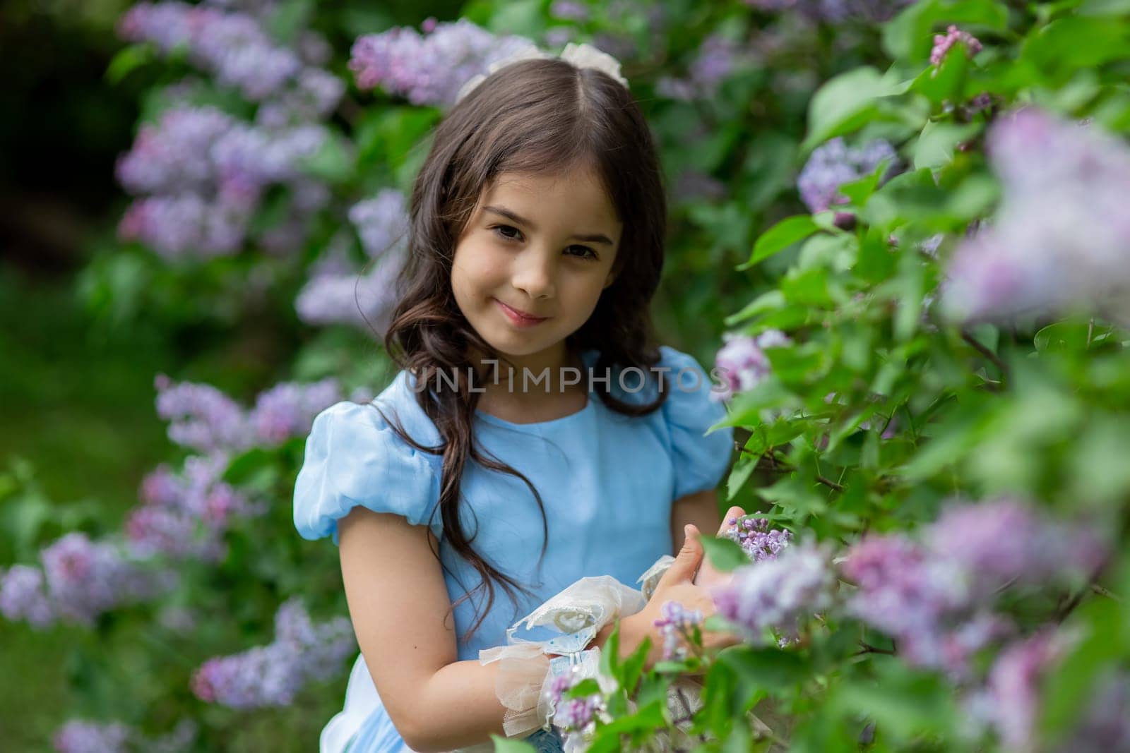 Young caucasian girl in blue dress smiling among lilac flowers in a garden.
