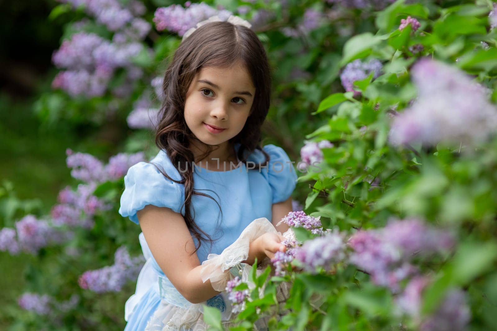 Young caucasian girl in blue dress among lilac blossoms in garden.