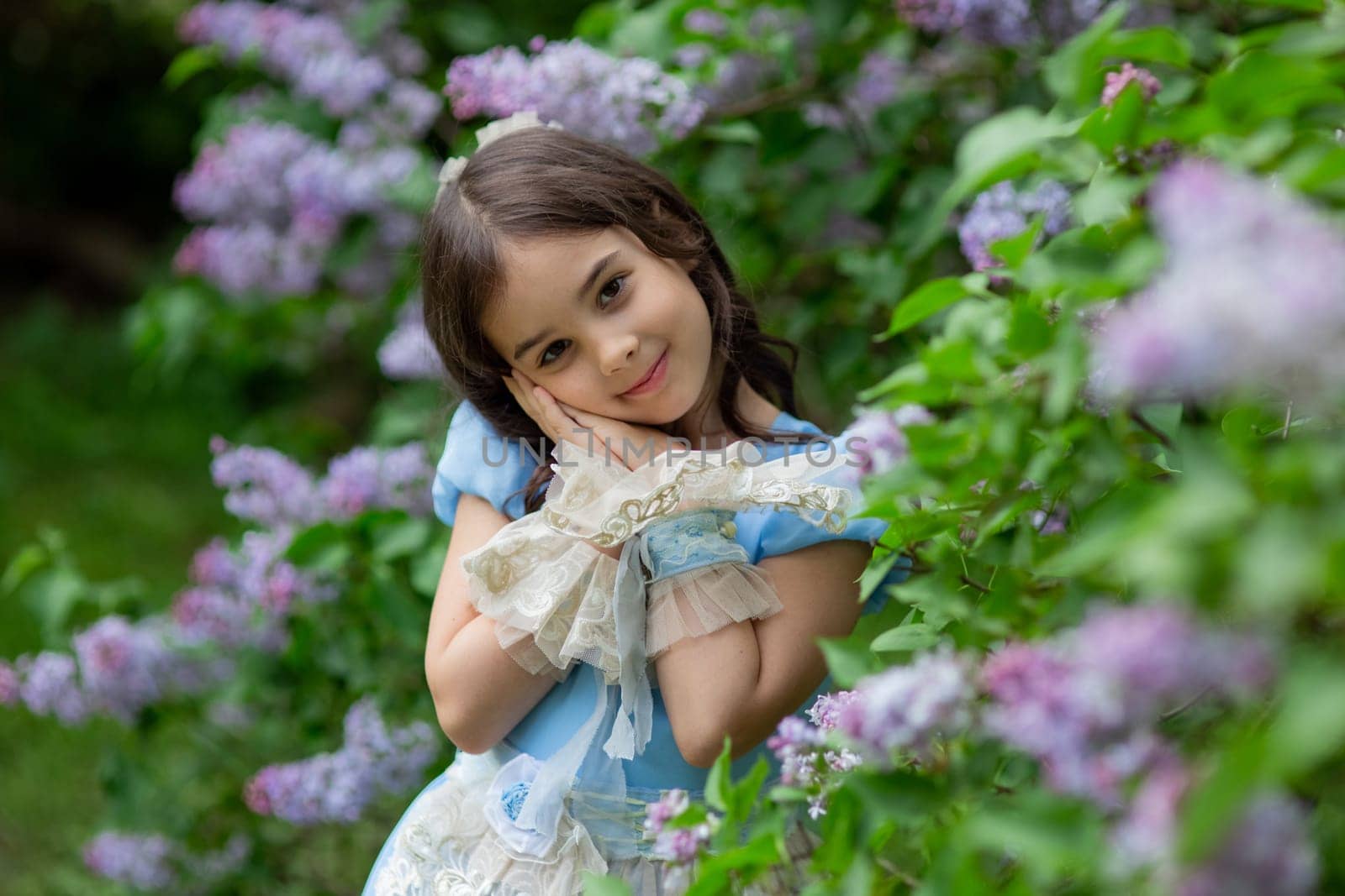 Young caucasian girl in blue dress among lilac flowers in garden.