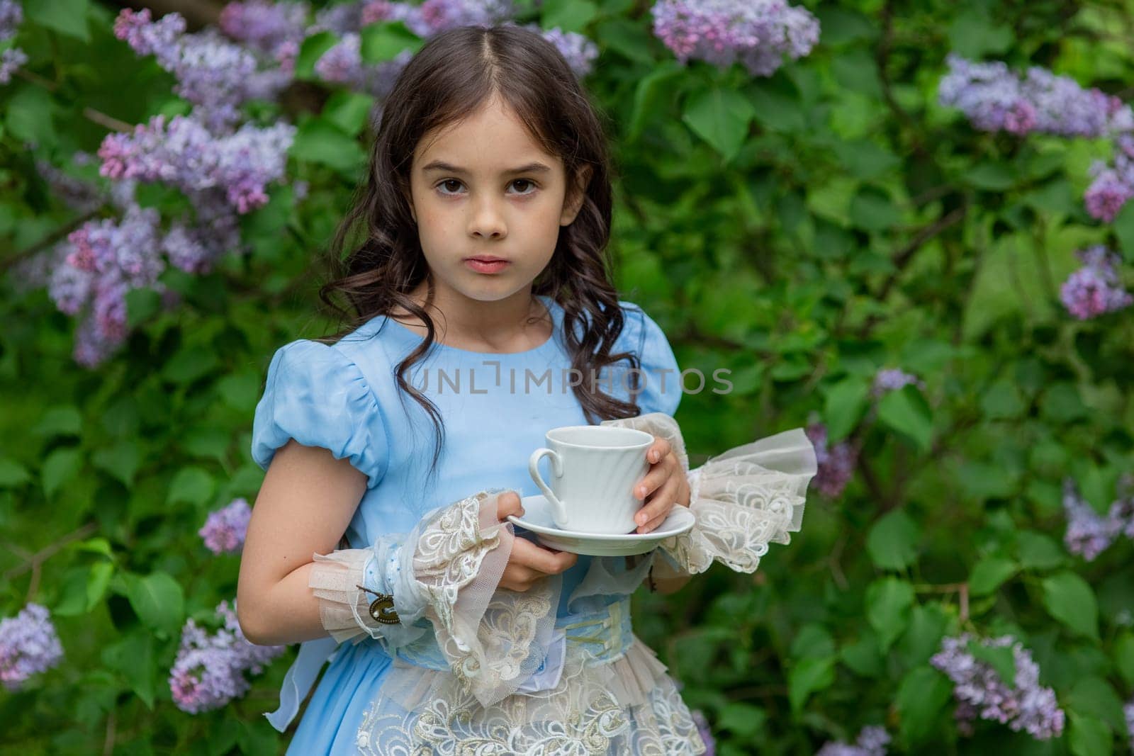 Young caucasian girl in blue dress holding teacup surrounded by lilac flowers.