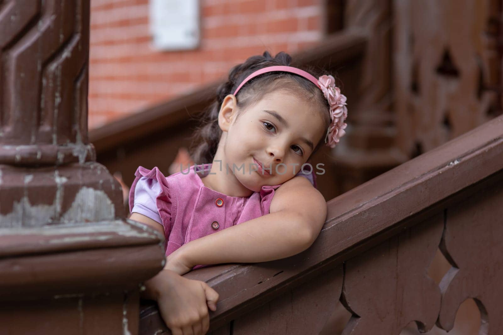 Hispanic young girl in pink outfit resting on wooden balcony railing.