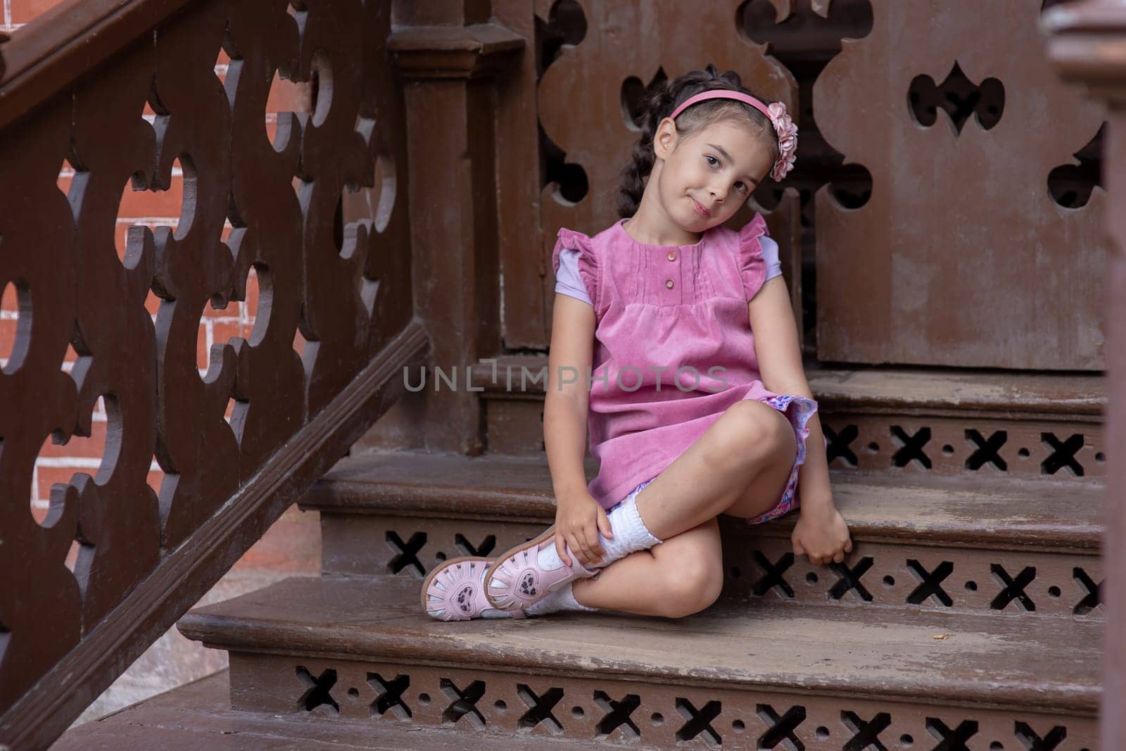 Young hispanic girl in pink dress sitting on ornate wooden stairs in thoughtful pose.