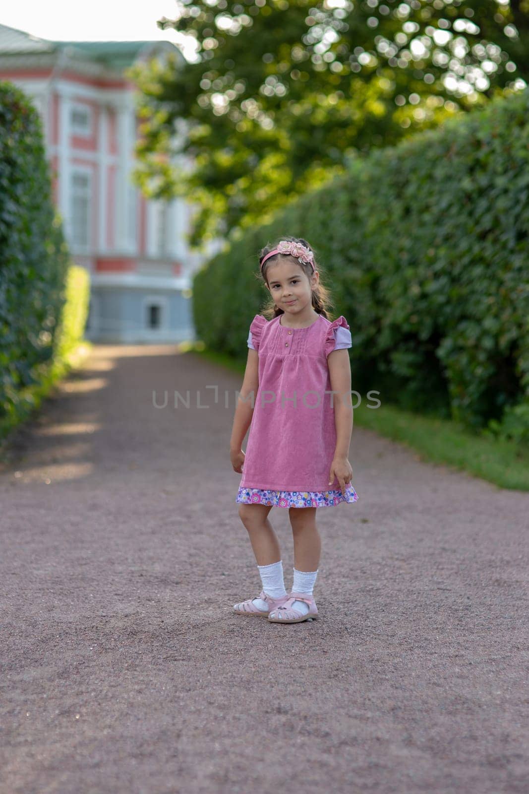 Young caucasian girl in pink dress smiling on garden path outdoors.