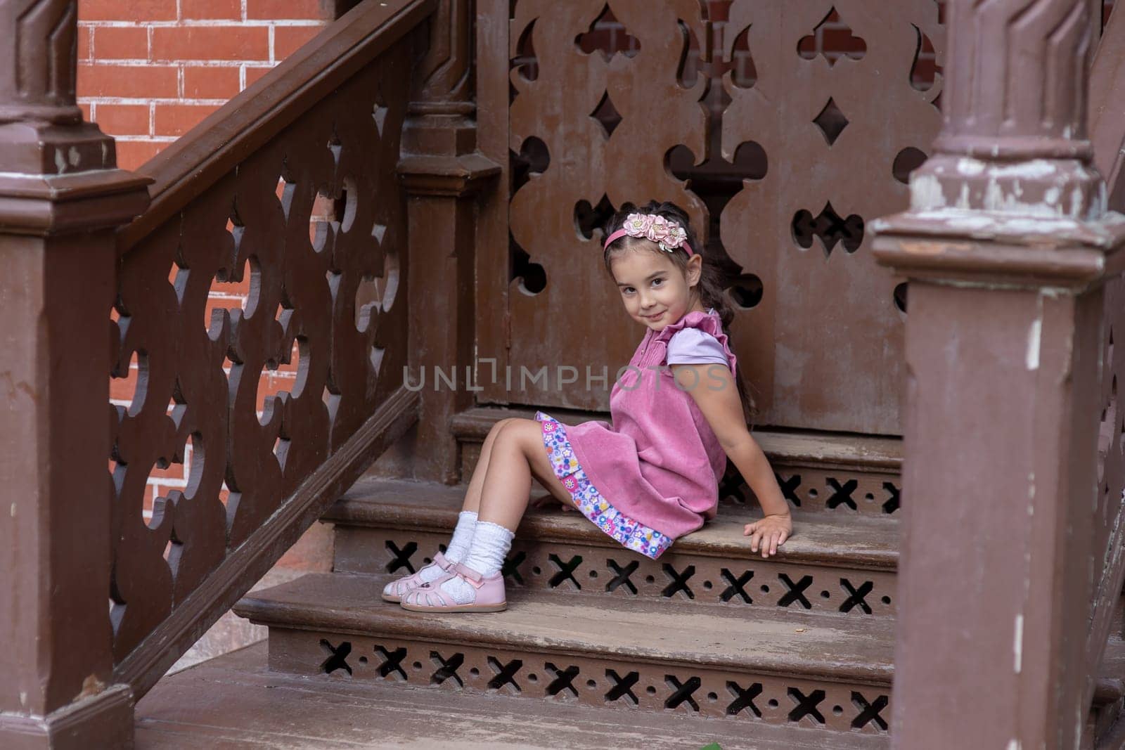 Young caucasian girl sitting on decorative wooden stairs in pink dress.