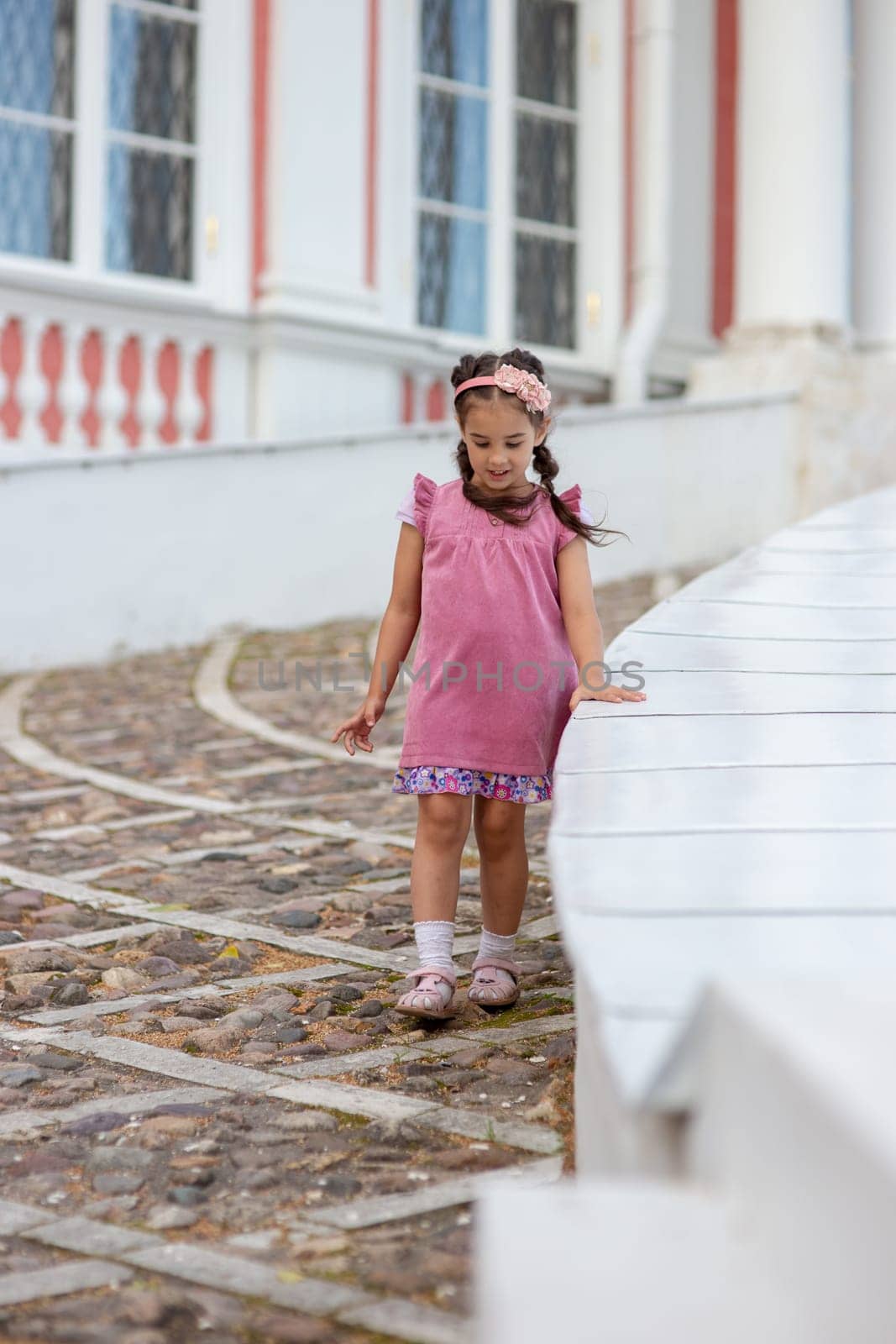 Young girl in pink dress walking on cobblestone path in front of vintage building.