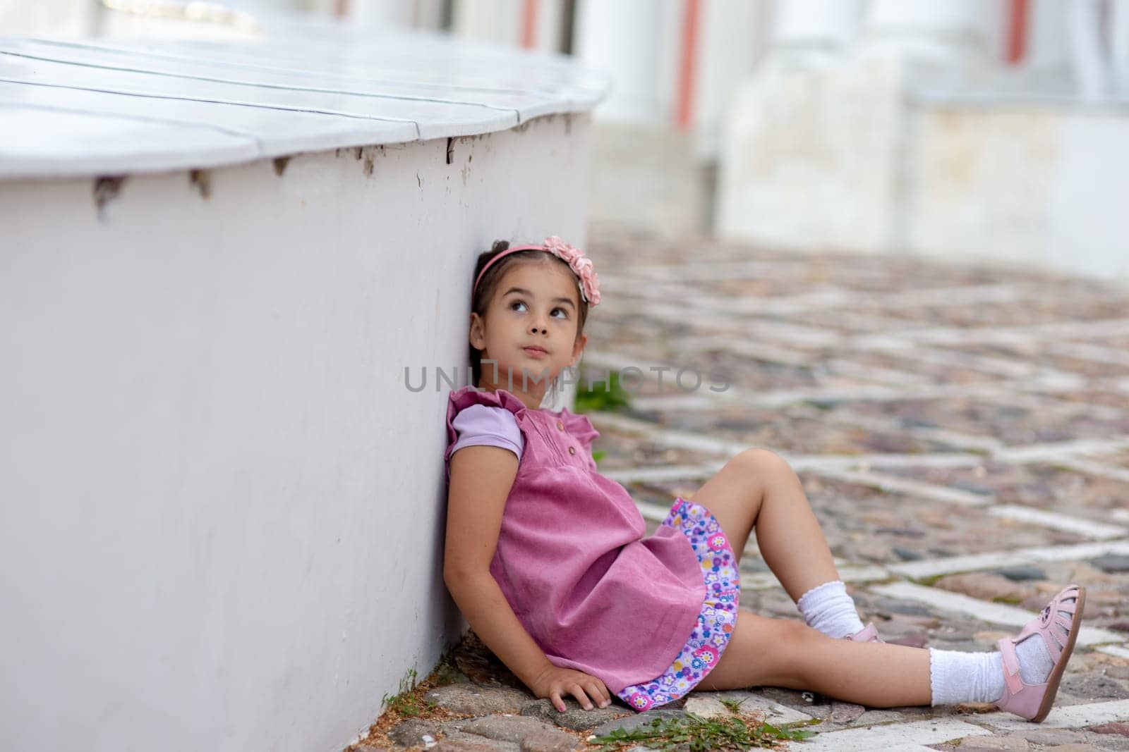 Young girl in pink dress sitting on cobblestone street.