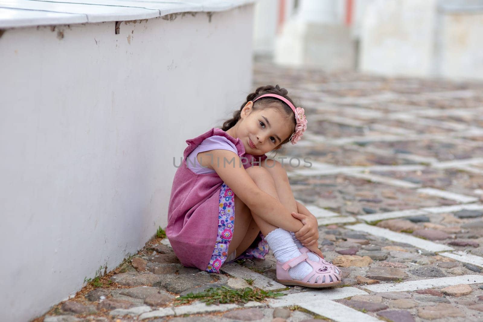 Young hispanic girl sitting on cobblestone street wearing pink dress and headband.