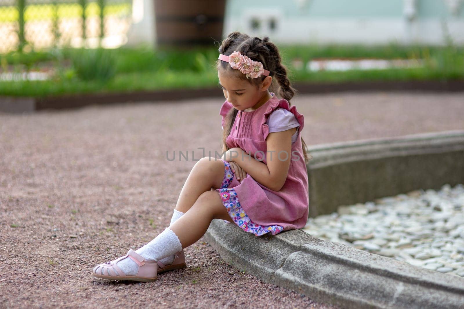 Young caucasian girl in pink dress sitting by garden fountain thoughtfully.