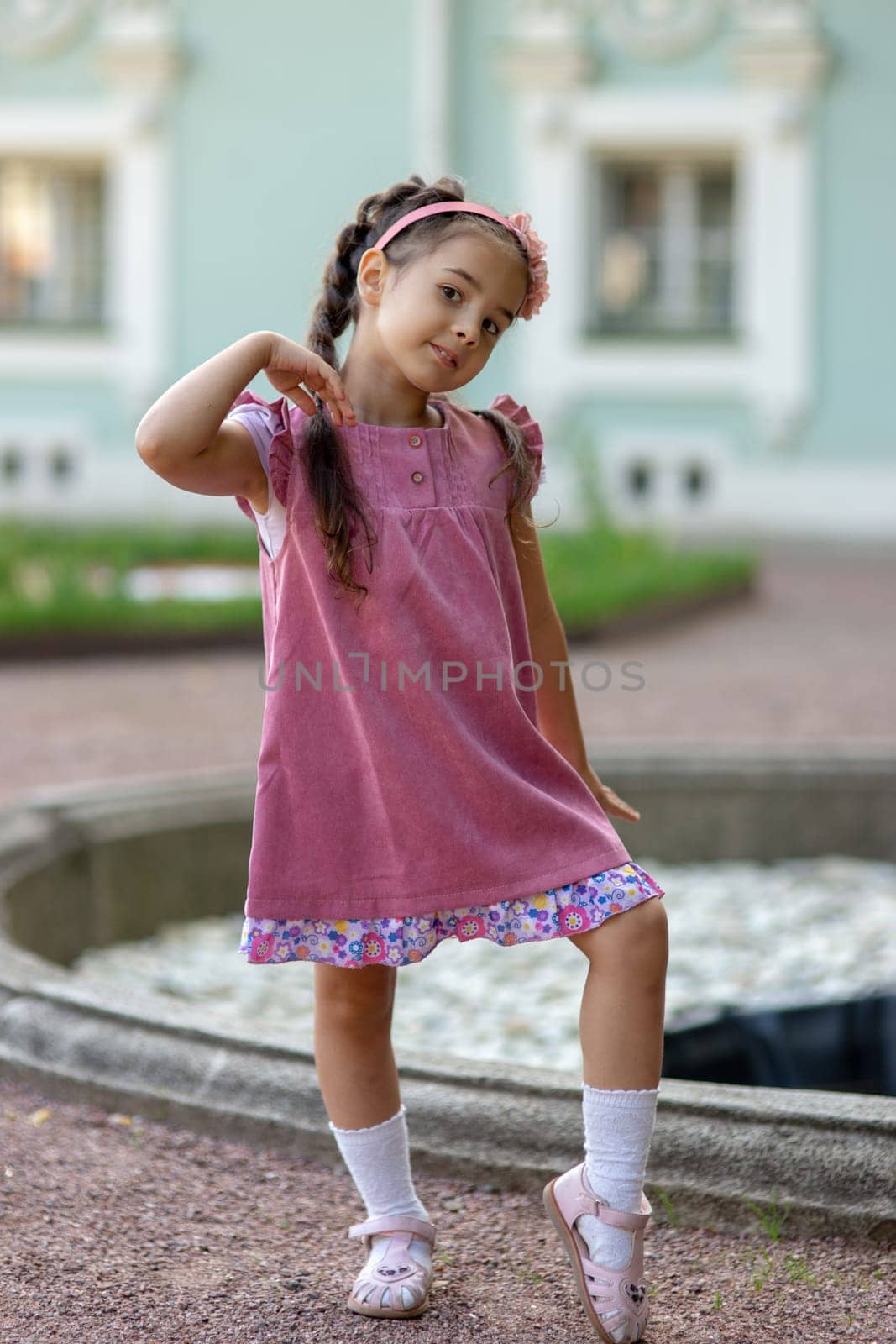 Young caucasian girl posing playfully in a pink dress near a fountain.