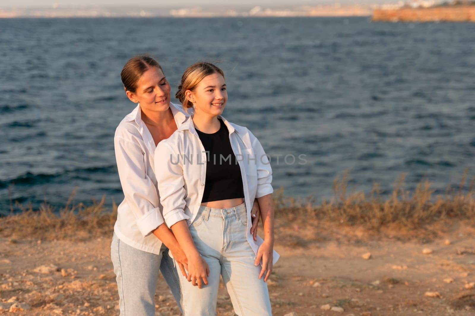 Two women, one wearing a white shirt and the other a black top, stand by a body of water, smiling and looking at each other.