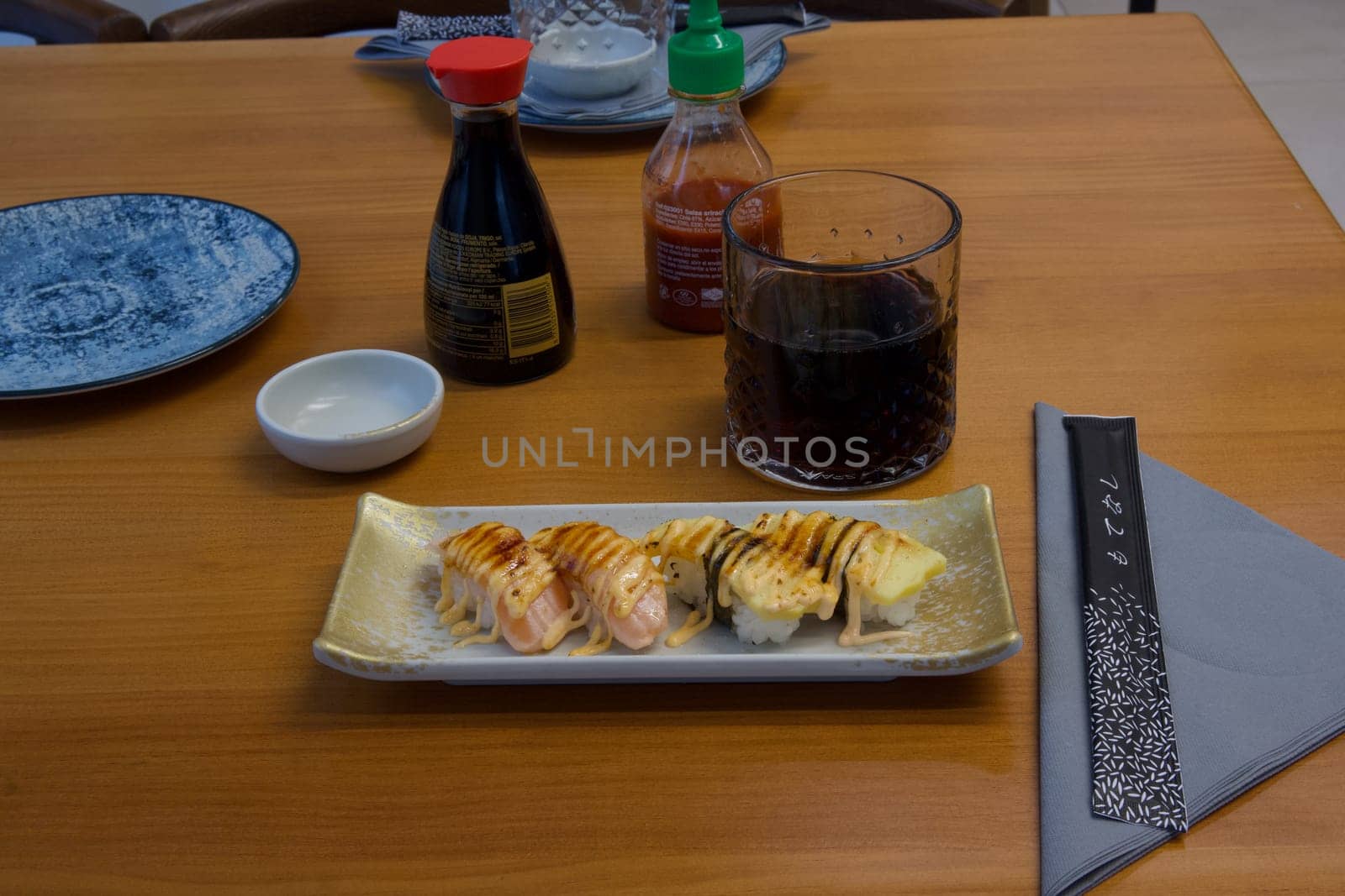 A plate of sushi sits on a wooden table with a napkin folded into a triangle. The table is set with condiments and a glass of wine. Scene is relaxed and inviting