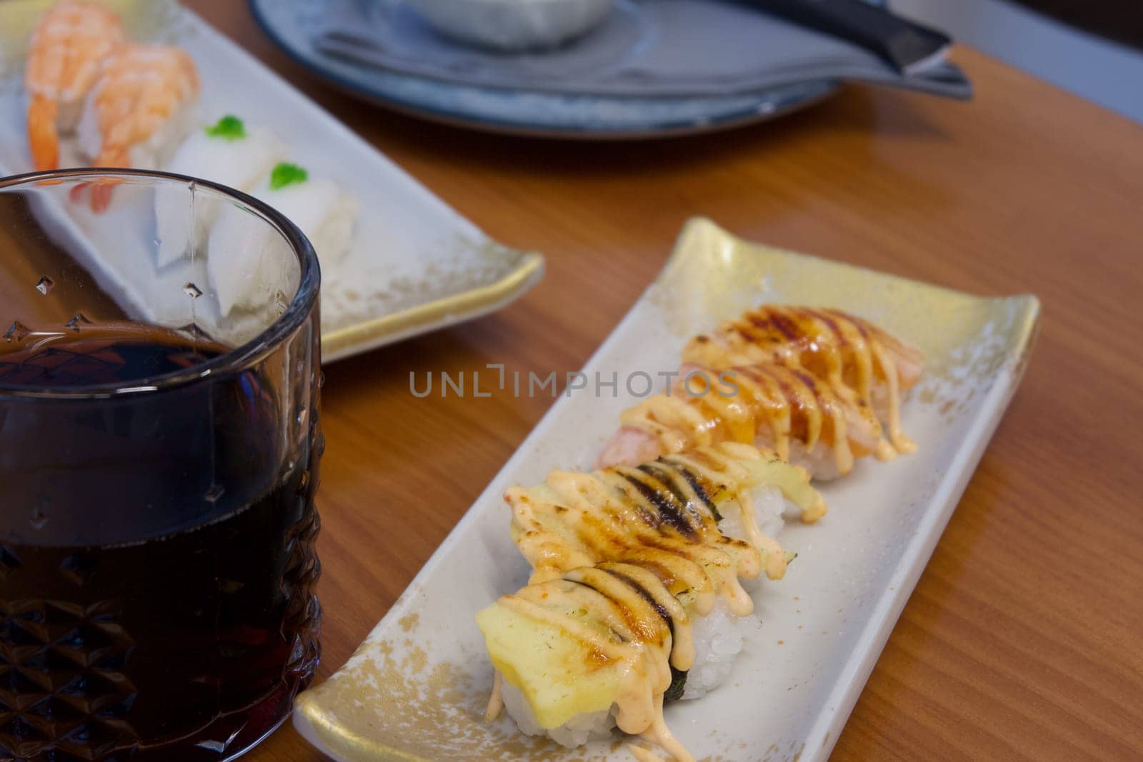 A plate of sushi with a sauce on it is placed on a wooden table next to a glass of soda. The sushi is arranged in a way that it looks appetizing and inviting. The glass of soda is half full
