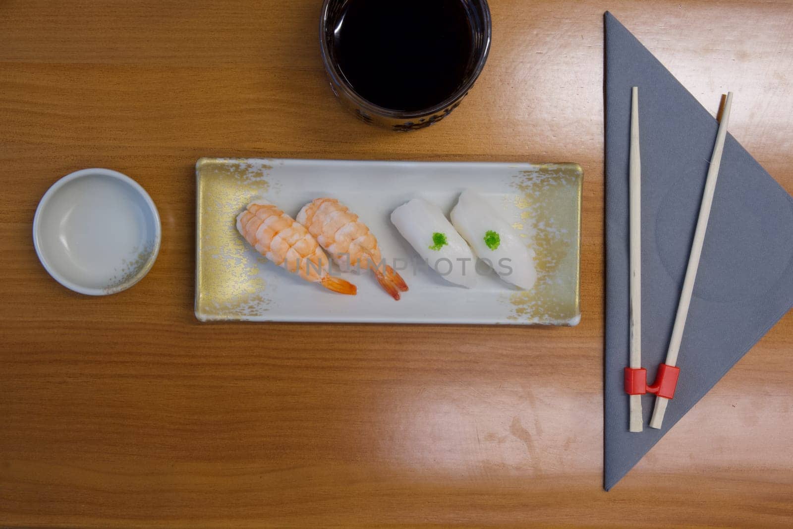 A plate of sushi with a shrimp and a white piece of fish on it. A cup of black coffee is on the table. A napkin is folded into a triangle and placed on the table