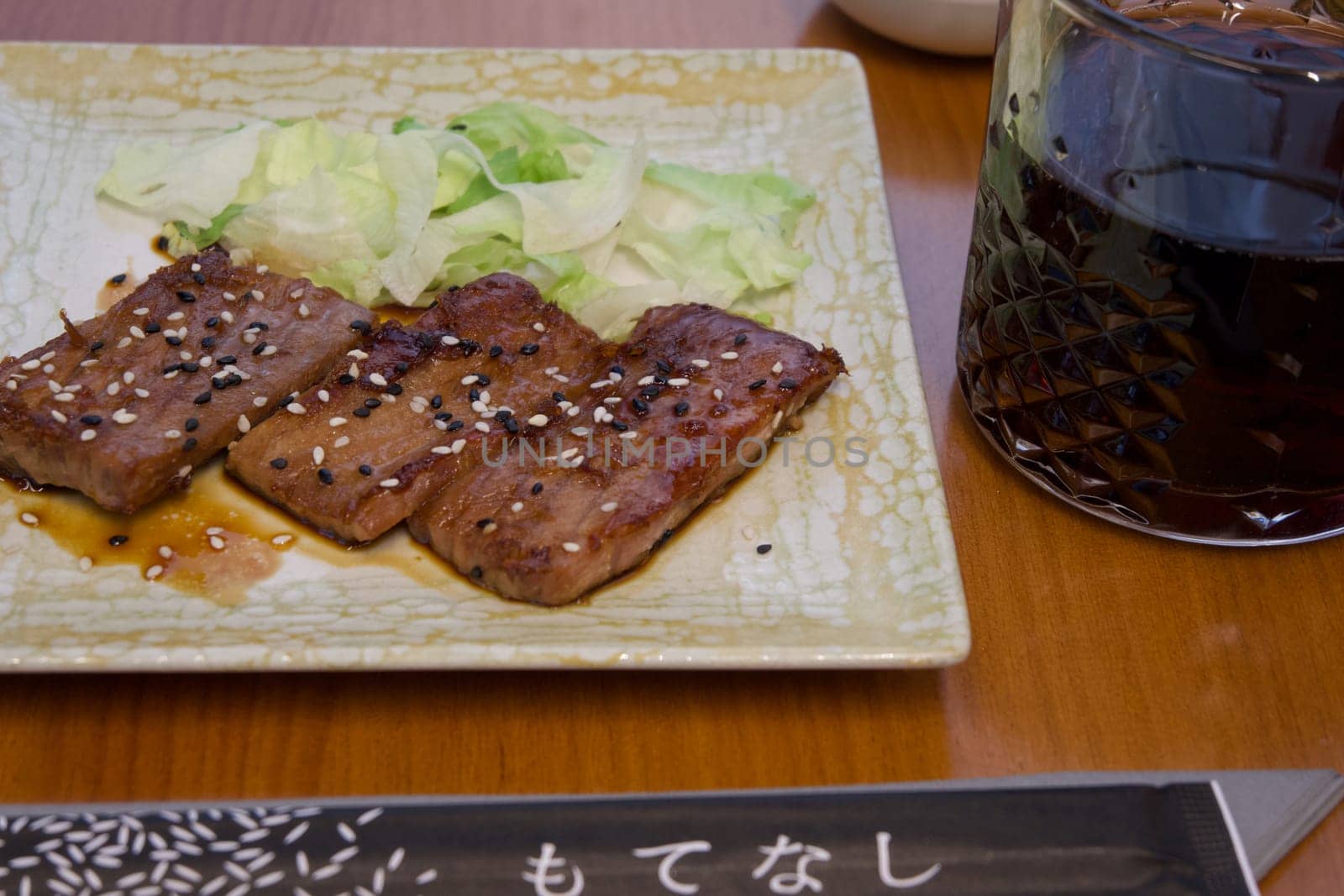 A plate of meat with sesame seeds and a glass of dark liquid. The plate is on a wooden table