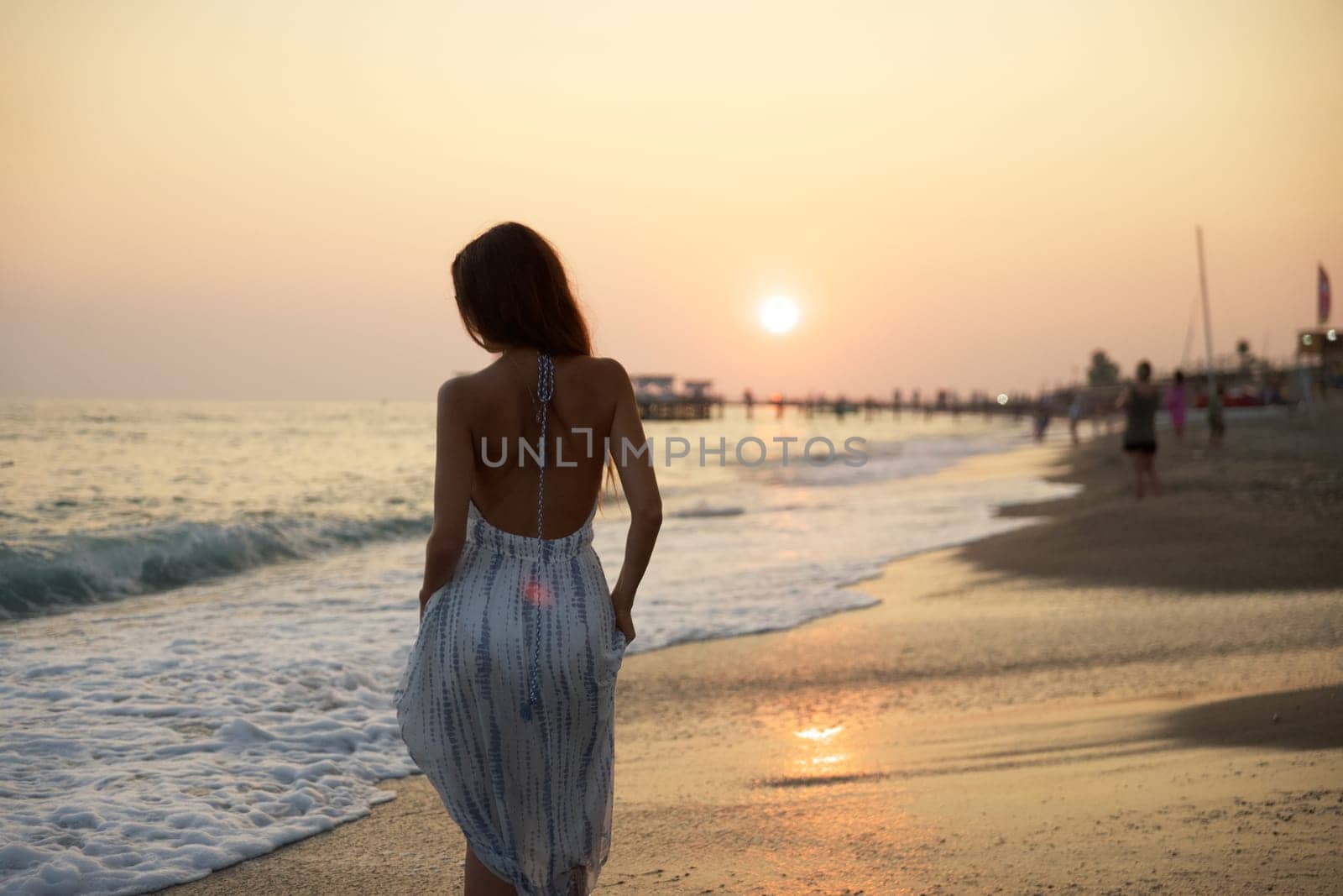 A woman in a flowing dress walks along a sunset beach, her back to the camera, with soft waves and a scenic pier in the distance, creating a peaceful and serene coastal atmosphere.