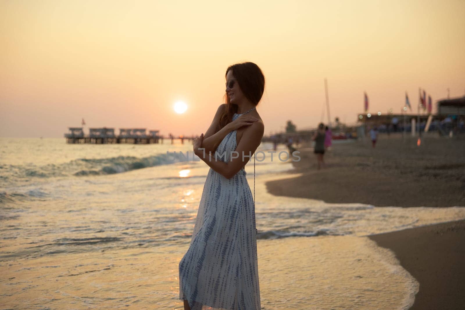 A peaceful sunset view of a woman walking barefoot on a sandy beach, surrounded by gentle waves, glowing skies, and a tranquil pier, creating a dreamy and relaxing seaside atmosphere.