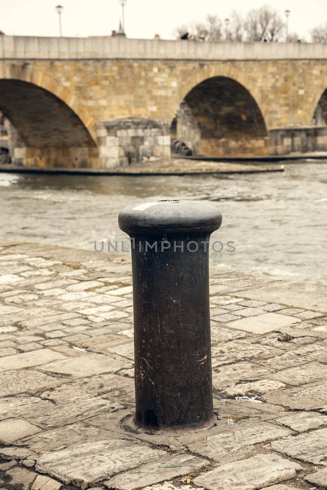 A worn mooring bollard on a historic cobblestone riverbank, set against the backdrop of an ancient stone bridge. Ideal for themes of history, travel, and urban heritage