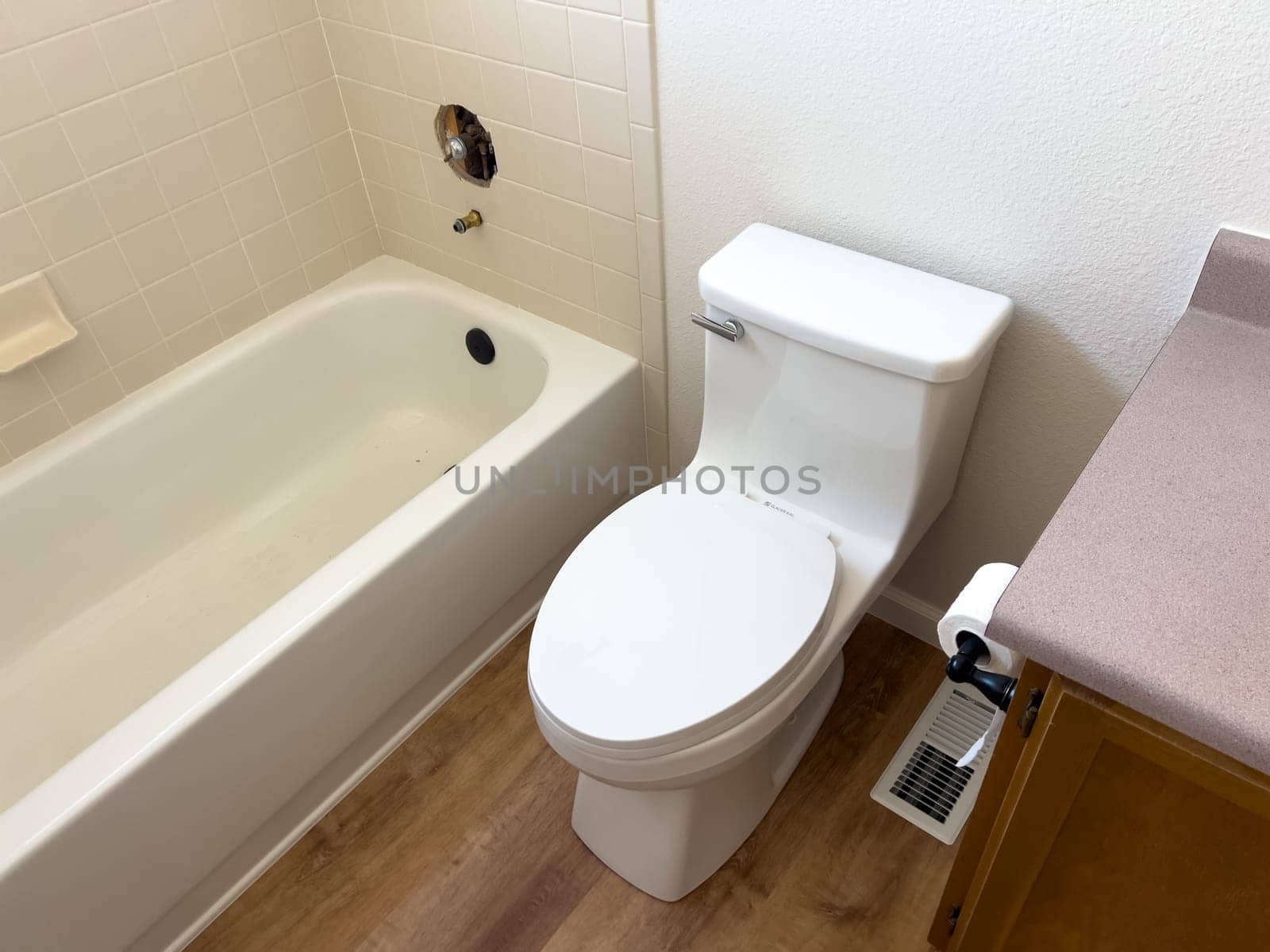 Broomfield, Colorado, USA-January 12, 2025-A clean modern white toilet installed in a bathroom next to a bathtub The wooden floor and beige countertop contrast with the white fixtures A toilet paper holder is mounted on the side of the vanity and an air vent is located on the floor near the toilet The bathtub has an open hole where a faucet or fixture is missing suggesting ongoing bathroom renovation or repair