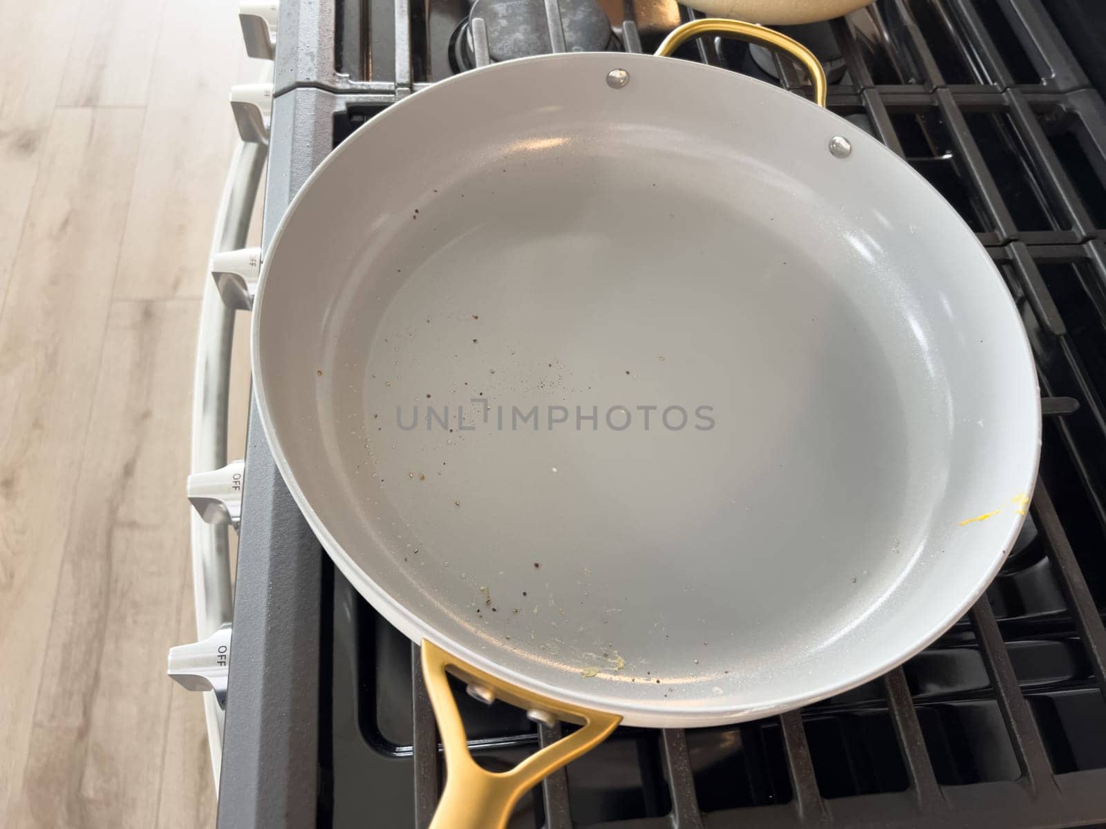 Castle Rock, Colorado, USA-January 17, 2024-A white ceramic pan with gold handles sits on a gas stove in a contemporary kitchen The stove features a stainless steel finish with multiple burners and control knobs A backsplash with white hexagonal tiles is visible in the background along with a utensil holder containing various kitchen tools and a bottle of cooking oil The scene showcases a clean and stylish cooking space