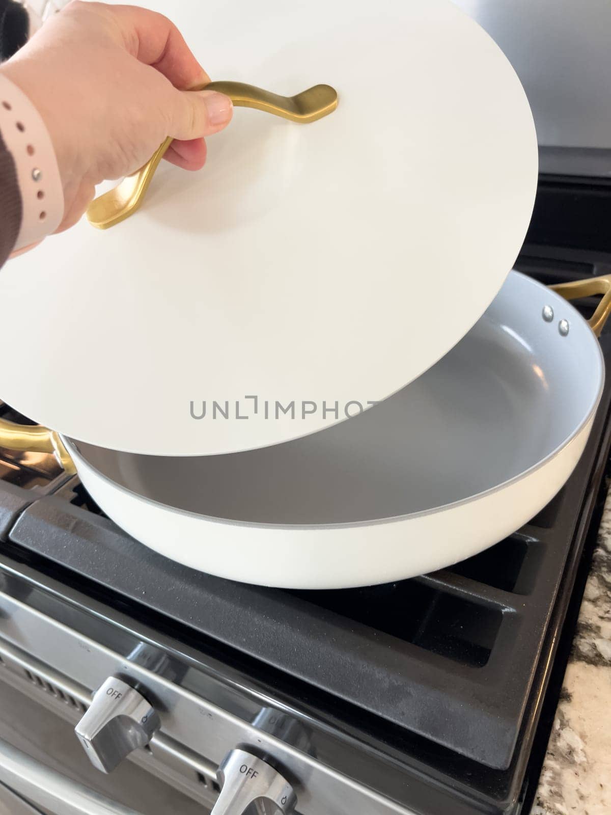 Castle Rock, Colorado, USA-January 17, 2024-A white ceramic pan with gold handles sits on a gas stove in a contemporary kitchen The stove features a stainless steel finish with multiple burners and control knobs A backsplash with white hexagonal tiles is visible in the background along with a utensil holder containing various kitchen tools and a bottle of cooking oil The scene showcases a clean and stylish cooking space