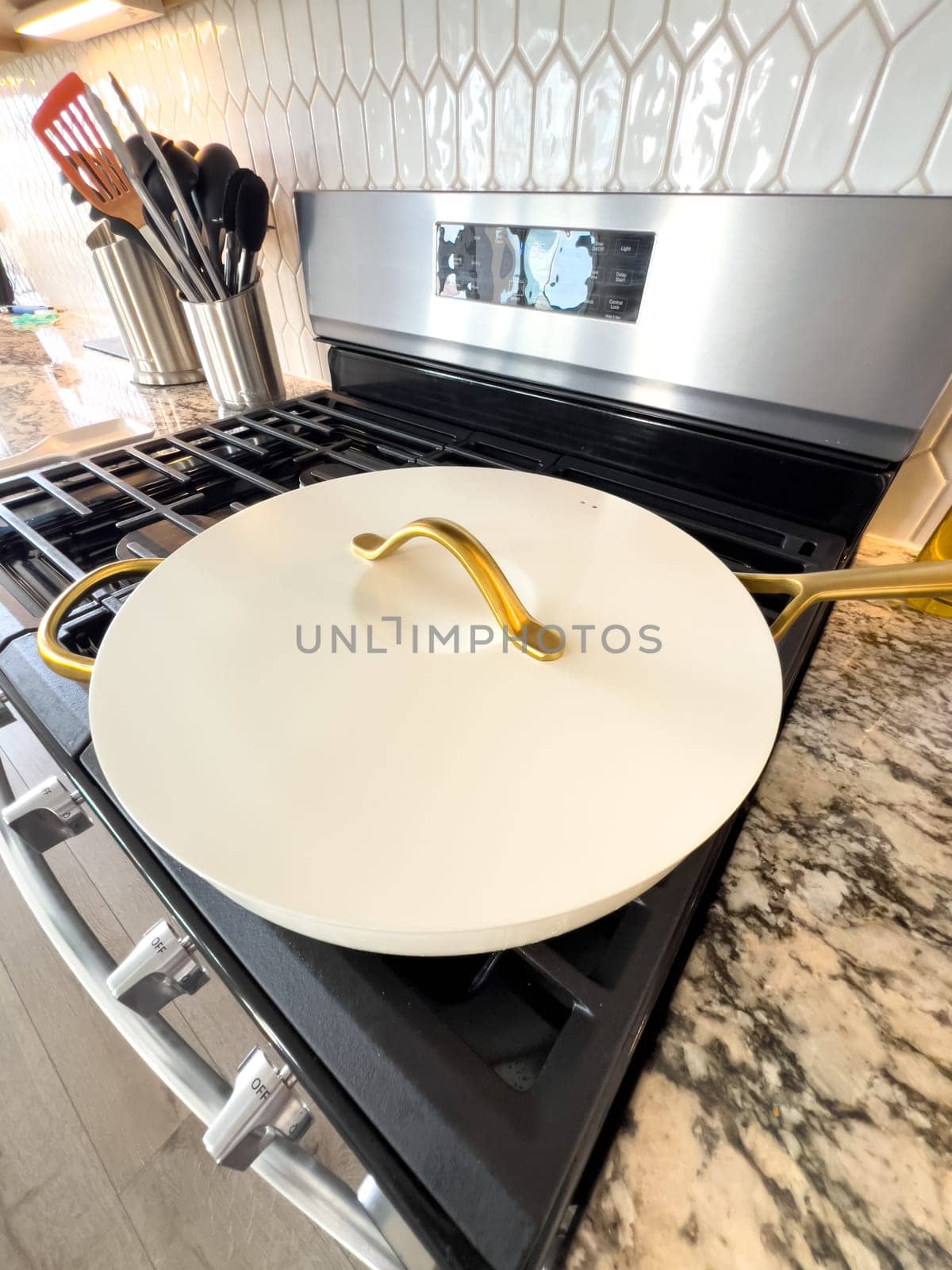 Castle Rock, Colorado, USA-January 17, 2024-A white ceramic pan with gold handles sits on a gas stove in a contemporary kitchen The stove features a stainless steel finish with multiple burners and control knobs A backsplash with white hexagonal tiles is visible in the background along with a utensil holder containing various kitchen tools and a bottle of cooking oil The scene showcases a clean and stylish cooking space