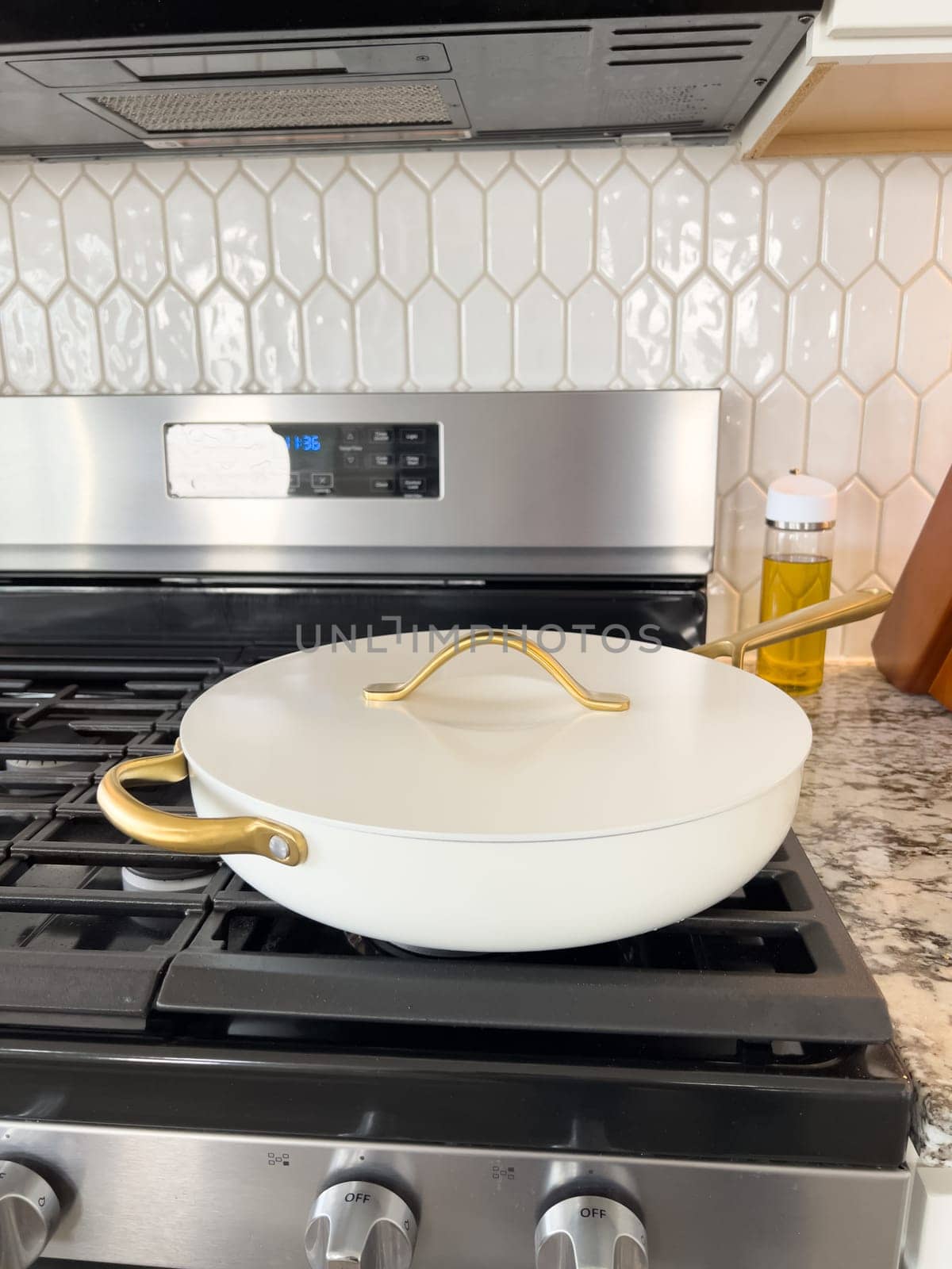 Castle Rock, Colorado, USA-January 17, 2024-A white ceramic pan with gold handles sits on a gas stove in a contemporary kitchen The stove features a stainless steel finish with multiple burners and control knobs A backsplash with white hexagonal tiles is visible in the background along with a utensil holder containing various kitchen tools and a bottle of cooking oil The scene showcases a clean and stylish cooking space