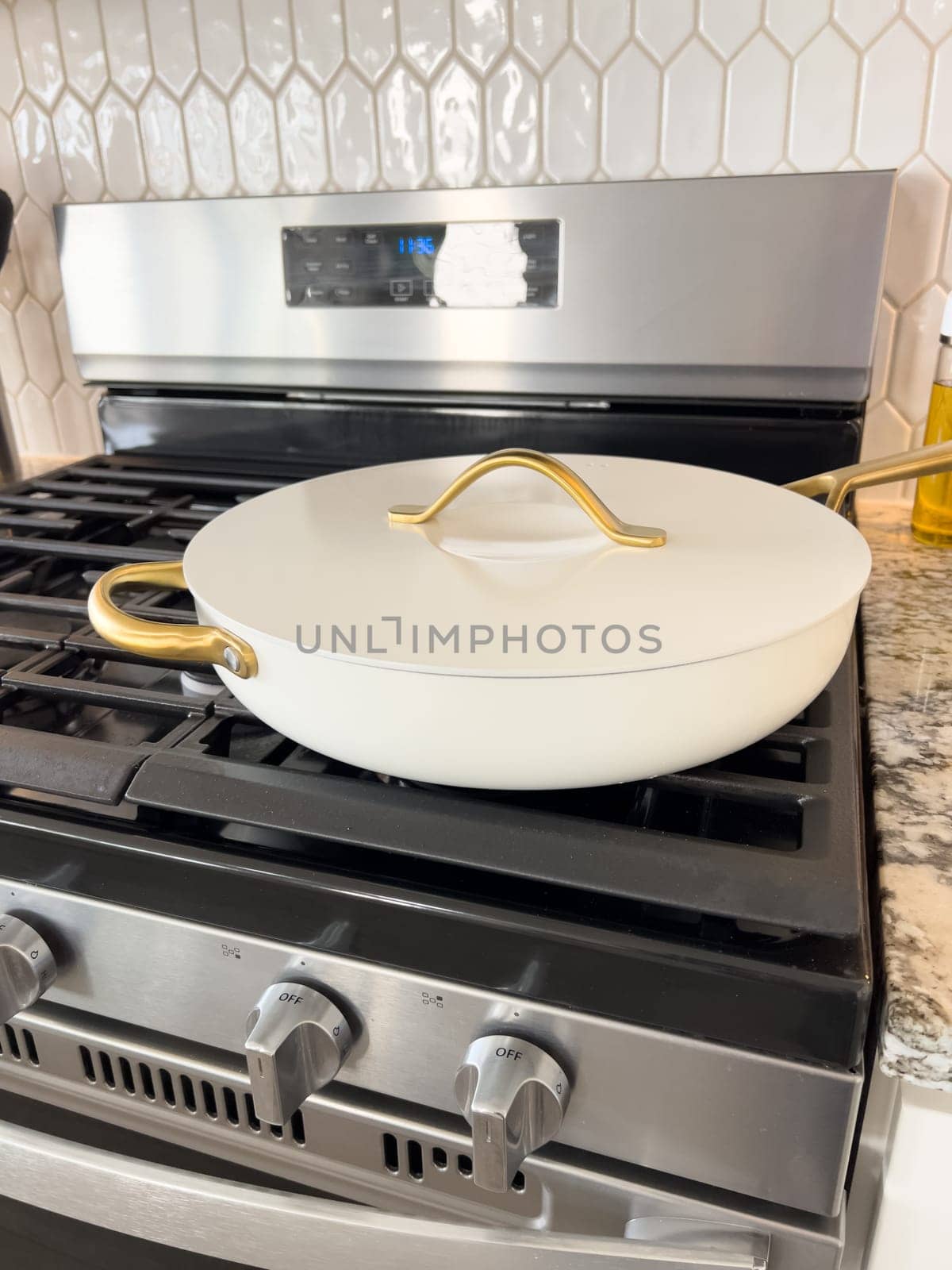 Castle Rock, Colorado, USA-January 17, 2024-A white ceramic pan with gold handles sits on a gas stove in a contemporary kitchen The stove features a stainless steel finish with multiple burners and control knobs A backsplash with white hexagonal tiles is visible in the background along with a utensil holder containing various kitchen tools and a bottle of cooking oil The scene showcases a clean and stylish cooking space