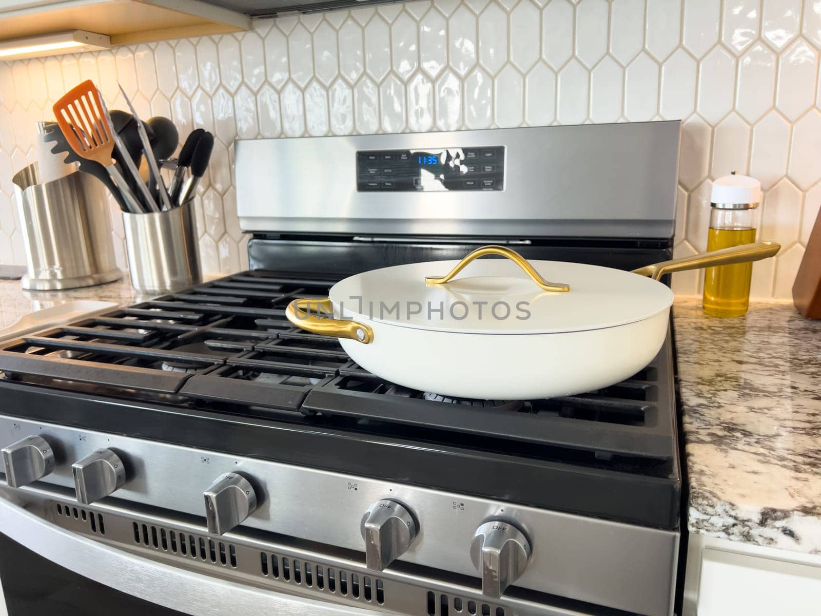 Castle Rock, Colorado, USA-January 17, 2024-A white ceramic pan with gold handles sits on a gas stove in a contemporary kitchen The stove features a stainless steel finish with multiple burners and control knobs A backsplash with white hexagonal tiles is visible in the background along with a utensil holder containing various kitchen tools and a bottle of cooking oil The scene showcases a clean and stylish cooking space