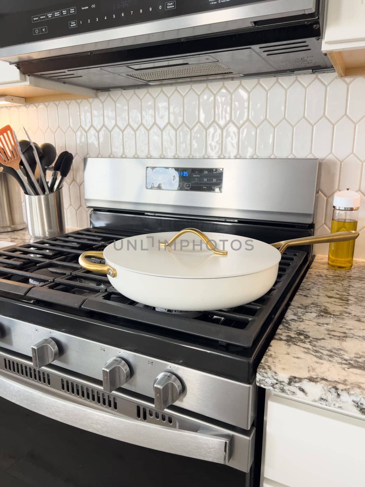 Castle Rock, Colorado, USA-January 17, 2024-A white ceramic pan with gold handles sits on a gas stove in a contemporary kitchen The stove features a stainless steel finish with multiple burners and control knobs A backsplash with white hexagonal tiles is visible in the background along with a utensil holder containing various kitchen tools and a bottle of cooking oil The scene showcases a clean and stylish cooking space