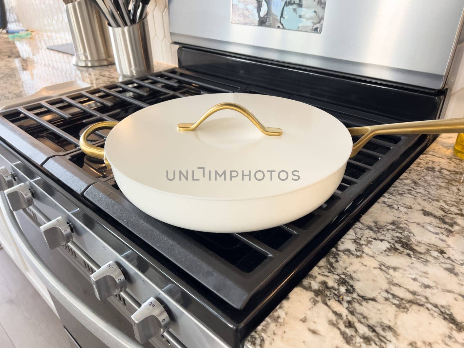 Castle Rock, Colorado, USA-January 17, 2024-A white ceramic pan with gold handles sits on a gas stove in a contemporary kitchen The stove features a stainless steel finish with multiple burners and control knobs A backsplash with white hexagonal tiles is visible in the background along with a utensil holder containing various kitchen tools and a bottle of cooking oil The scene showcases a clean and stylish cooking space