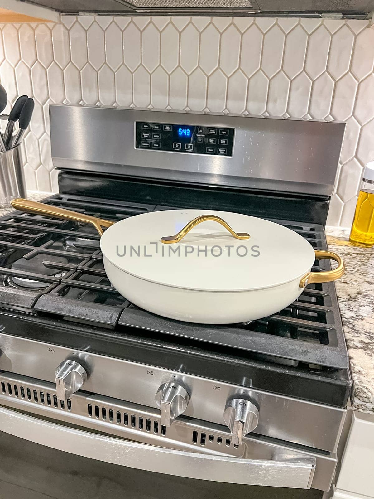 Castle Rock, Colorado, USA-January 17, 2024-A white ceramic pan with gold handles sits on a gas stove in a contemporary kitchen The stove features a stainless steel finish with multiple burners and control knobs A backsplash with white hexagonal tiles is visible in the background along with a utensil holder containing various kitchen tools and a bottle of cooking oil The scene showcases a clean and stylish cooking space